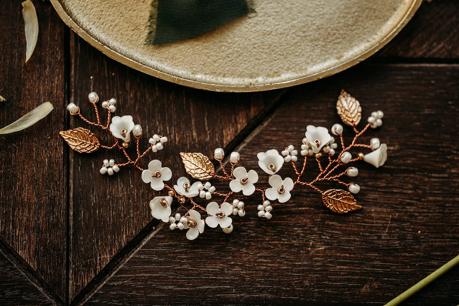 Eine dekorative Haarspange mit weißen Blüten, goldenen Blättern und Perlen auf einem dunklen Holztisch neben einem beige farbenen Teller.