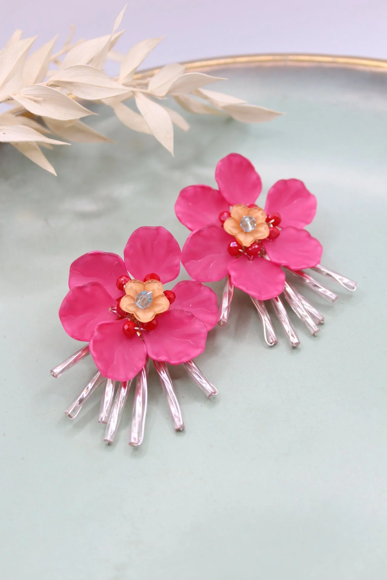 Zwei pinke Blumen-Ohrringe mit kleinen Kristallen, roten und gelben Akzenten, liegen auf einem hellblauen Untergrund, im Hintergrund ist getrocknetes weißes Gras.