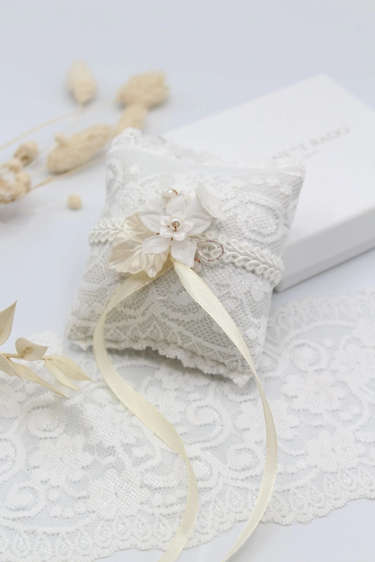 Weiße Hochzeitsoder Schmuck-Schachtel auf einem Spitzenstoff mit Blumen, Schleifen und Perlen, daneben ein weißes Buch.