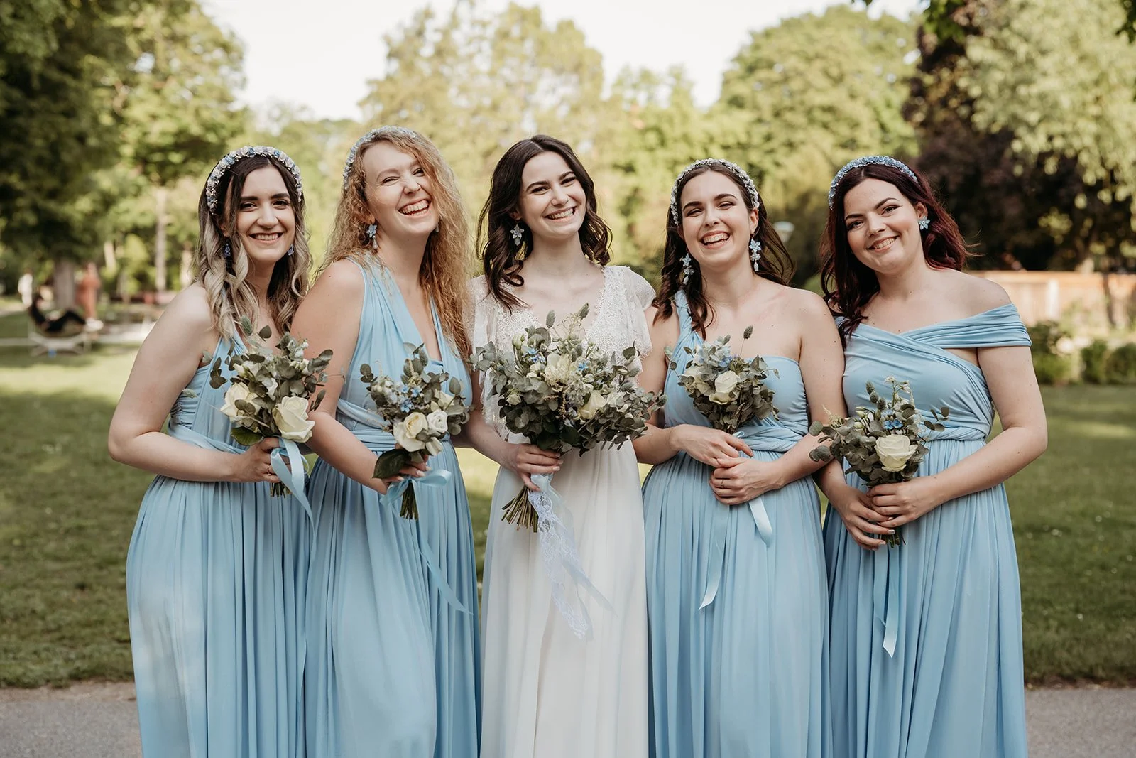 Fünf Frauen bei einer Hochzeit, vier Bräute in blauen Kleidern, eine Brautmutter in weiß, alle halten Blumensträuße und lachen in einem Park.