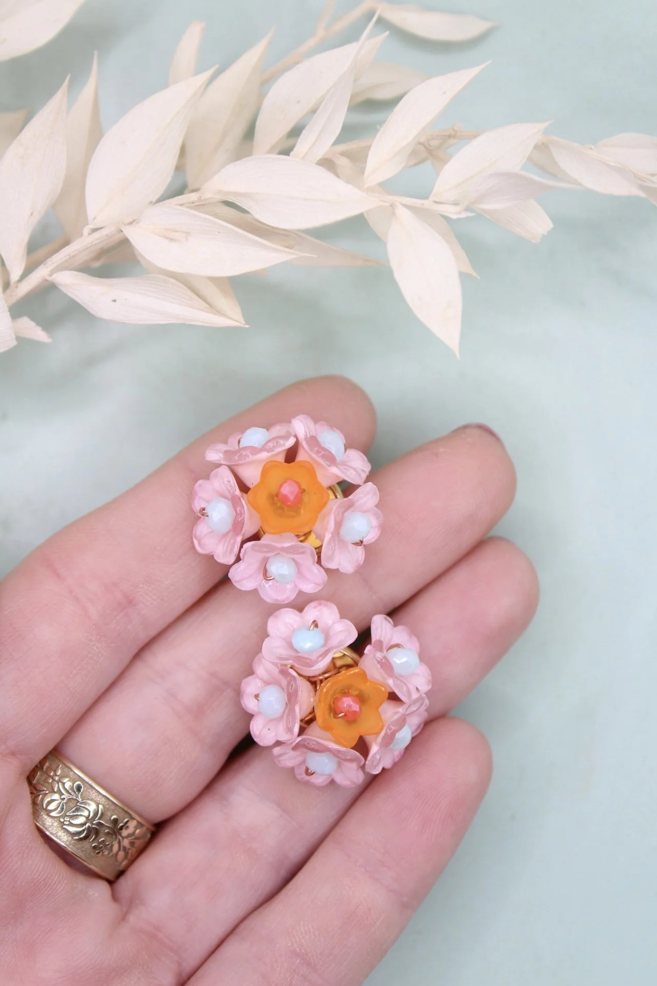 Nahaufnahme von einem Finger mit zwei pinken, blumenförmigen Ohrringen mit weißen Perlen und orangefarbenen Staubkörnern. Im Hintergrund eine getrocknete, weiße Staude auf blassem Hintergrund.