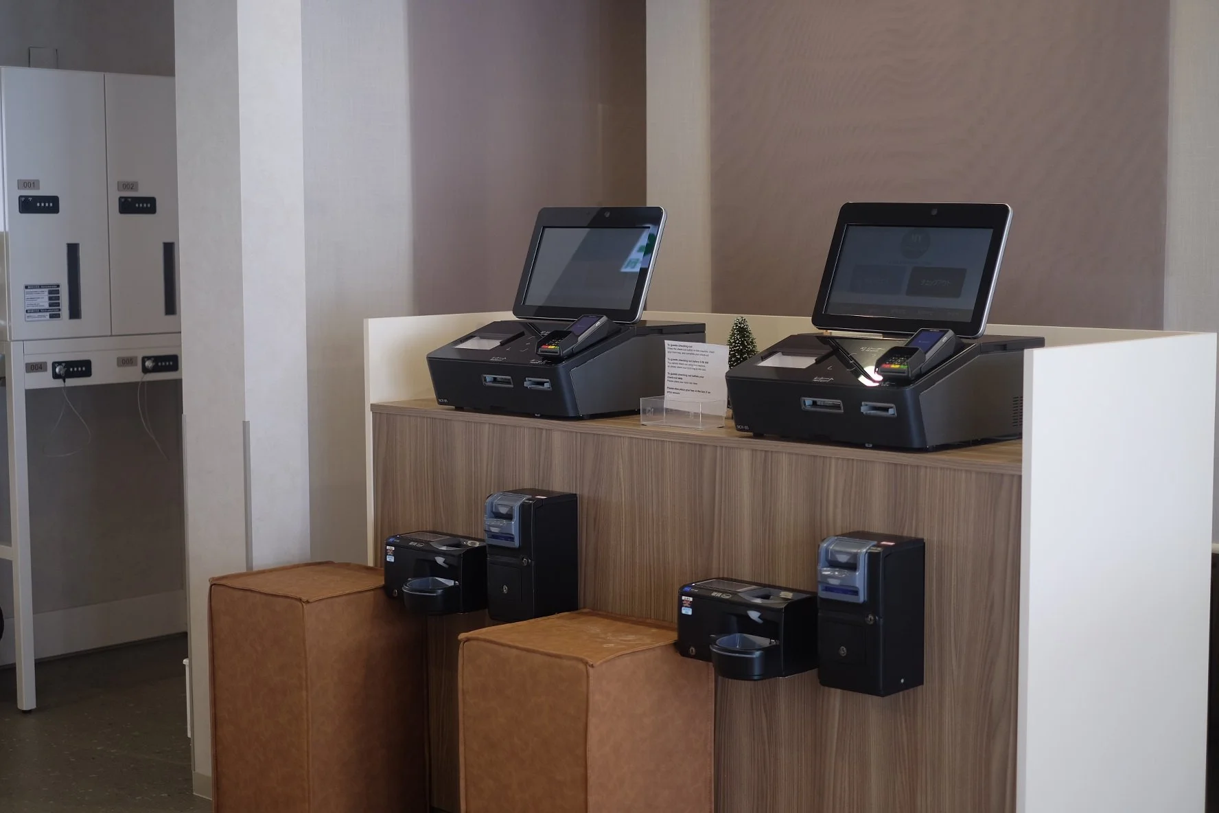 Two self-service checkout terminals with screens and card readers on a wooden counter. To the left are lockers, and below the counters are small print stations. The area is indoors, possibly in a retail store.