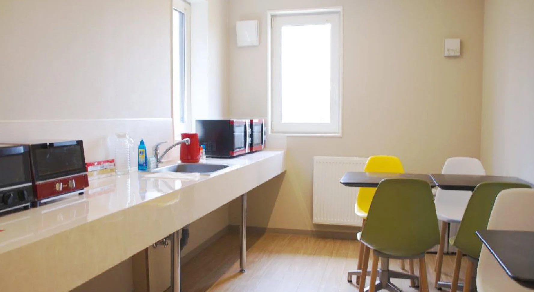 Kitchen with three toasters, a sink, microwave oven, and colorful chairs near a table by the window.