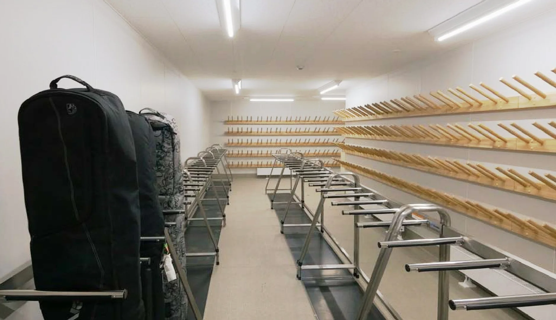Empty golf club storage room with rows of wall-mounted wooden racks for golf club hooks, several golf bags on trolleys, and gray flooring.