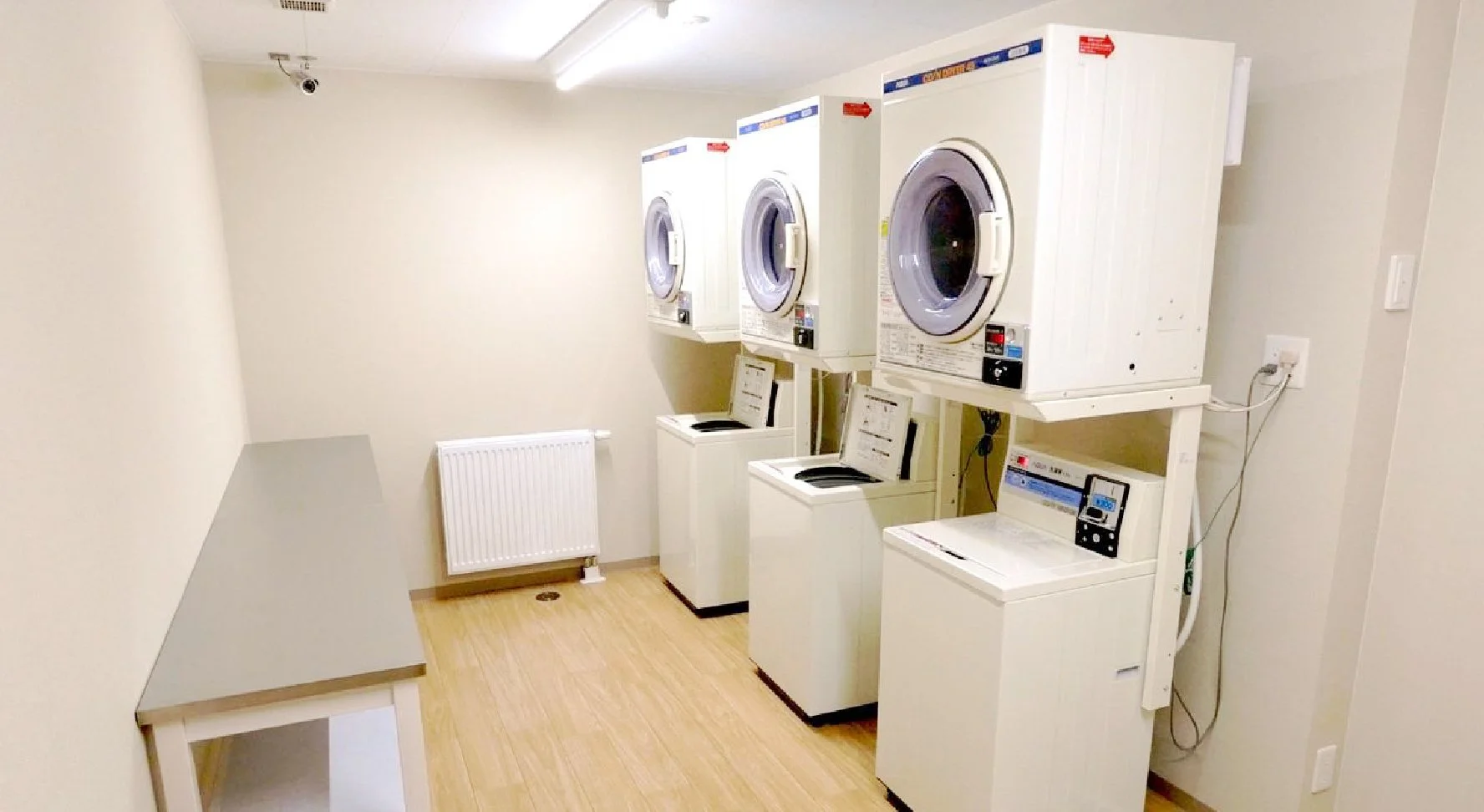 A small laundry room with three stacked white washing machines and dryers, a white radiator, and a gray table in the corner.