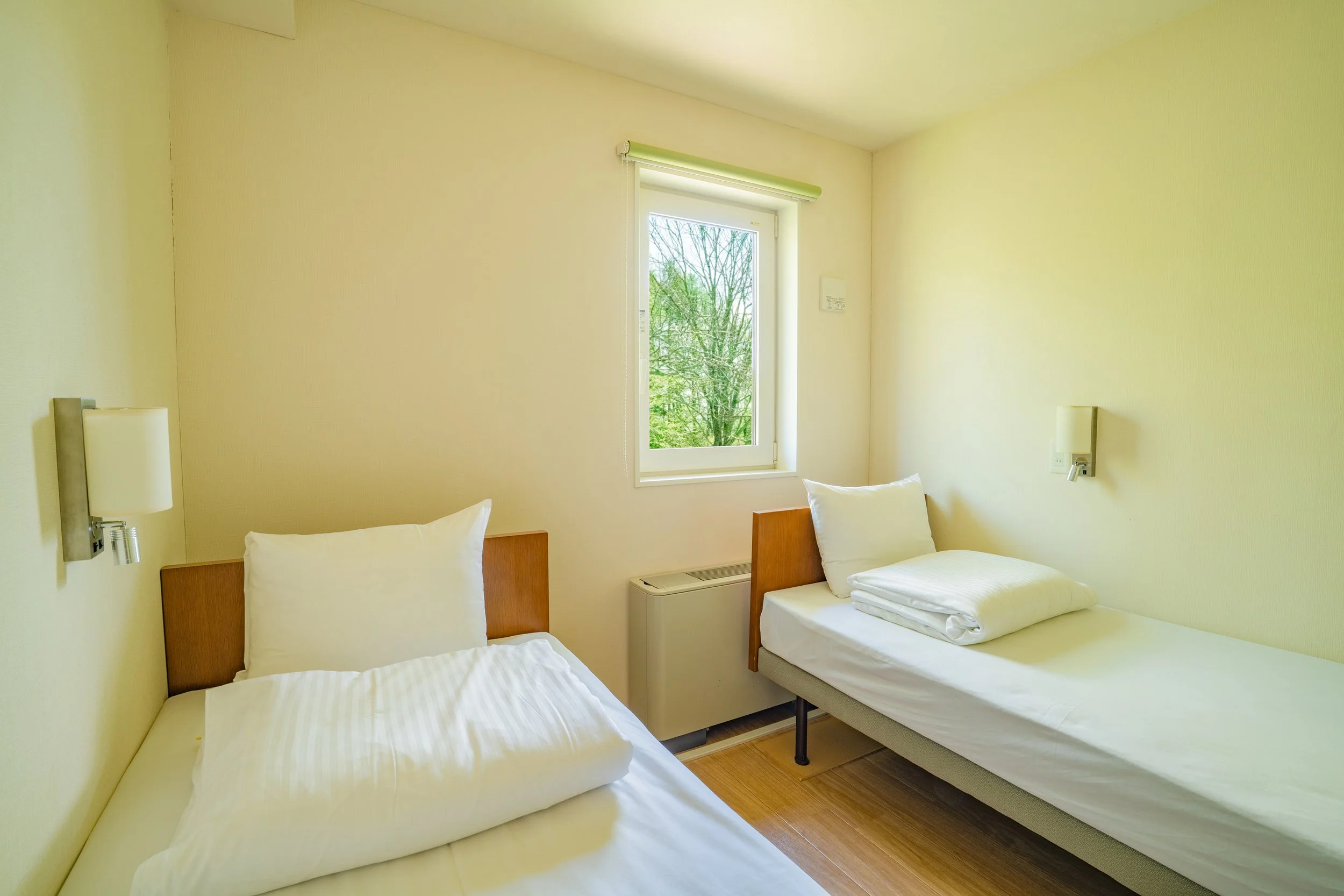 A hotel room with two single beds, each with white linens and pillows, a window with a view of trees outside, and modern wall-mounted lights.