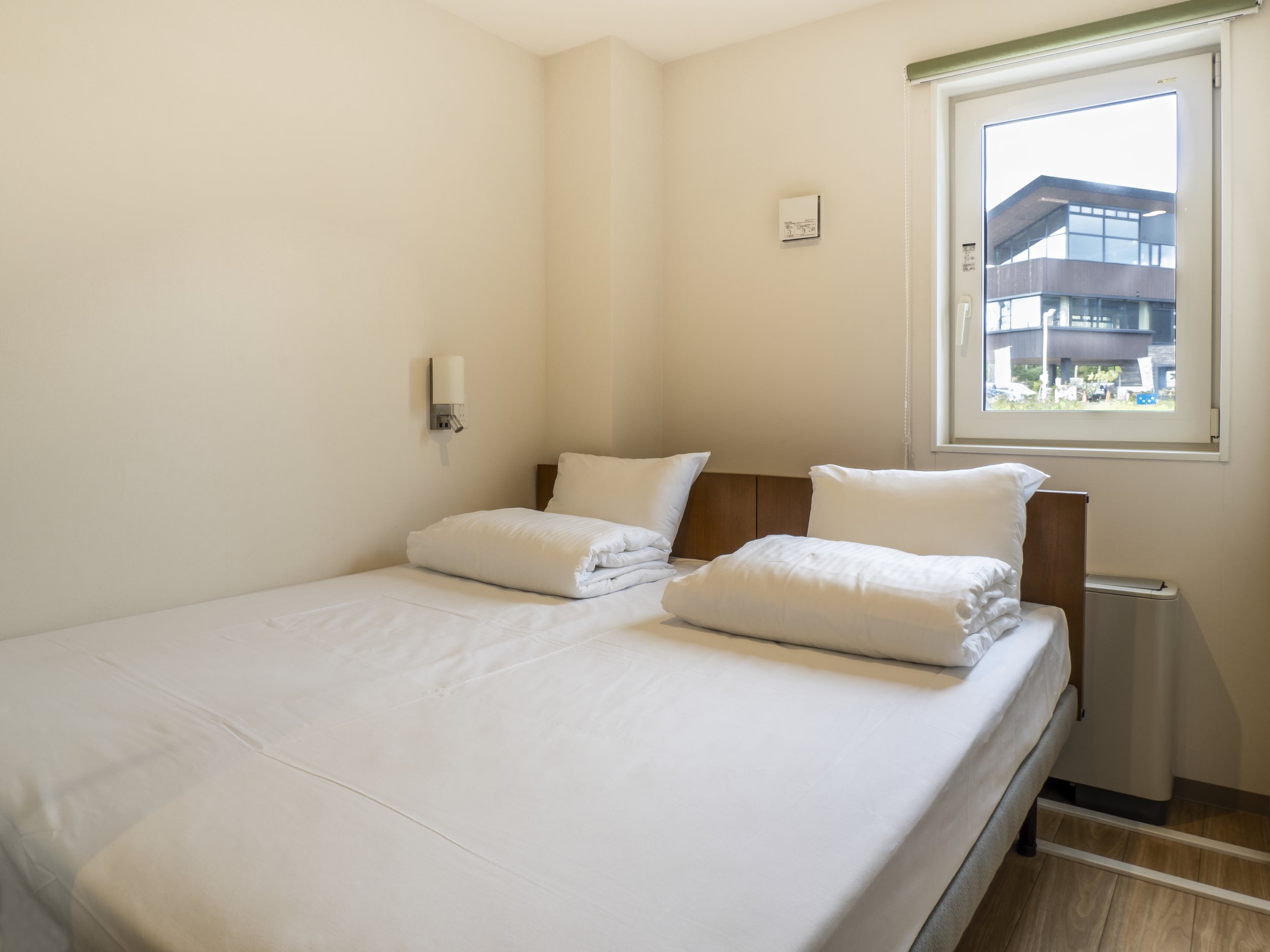 A hotel room with a bed made with white linens, two pillows, and a wooden headboard. There is a window with a view of a modern building outside, and a small nightstand on the right side of the bed.