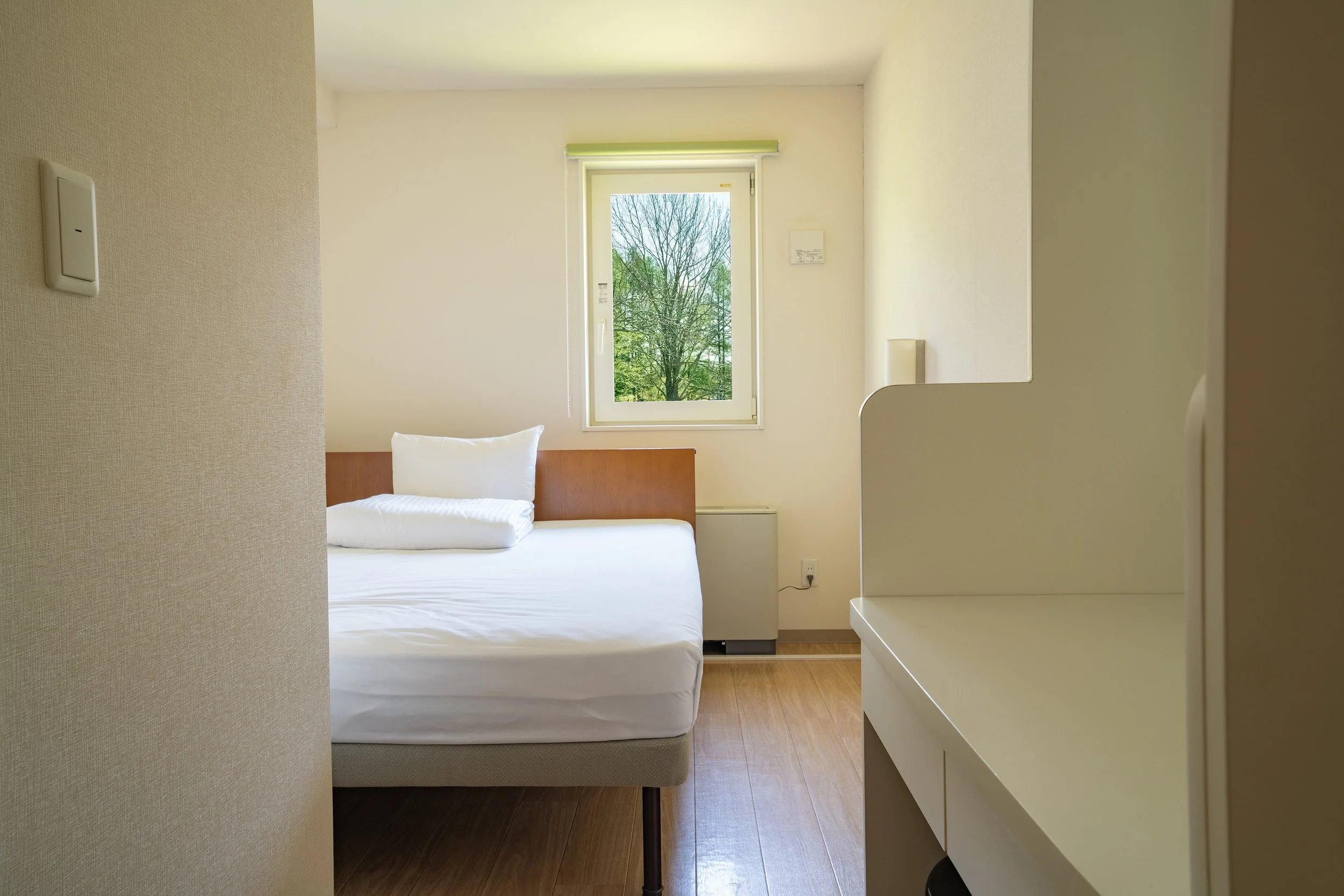 A minimalist hotel room with a bed near a window, beige walls, and wooden flooring. The bed has a white mattress and pillow, and there is a white shelf on the right side.