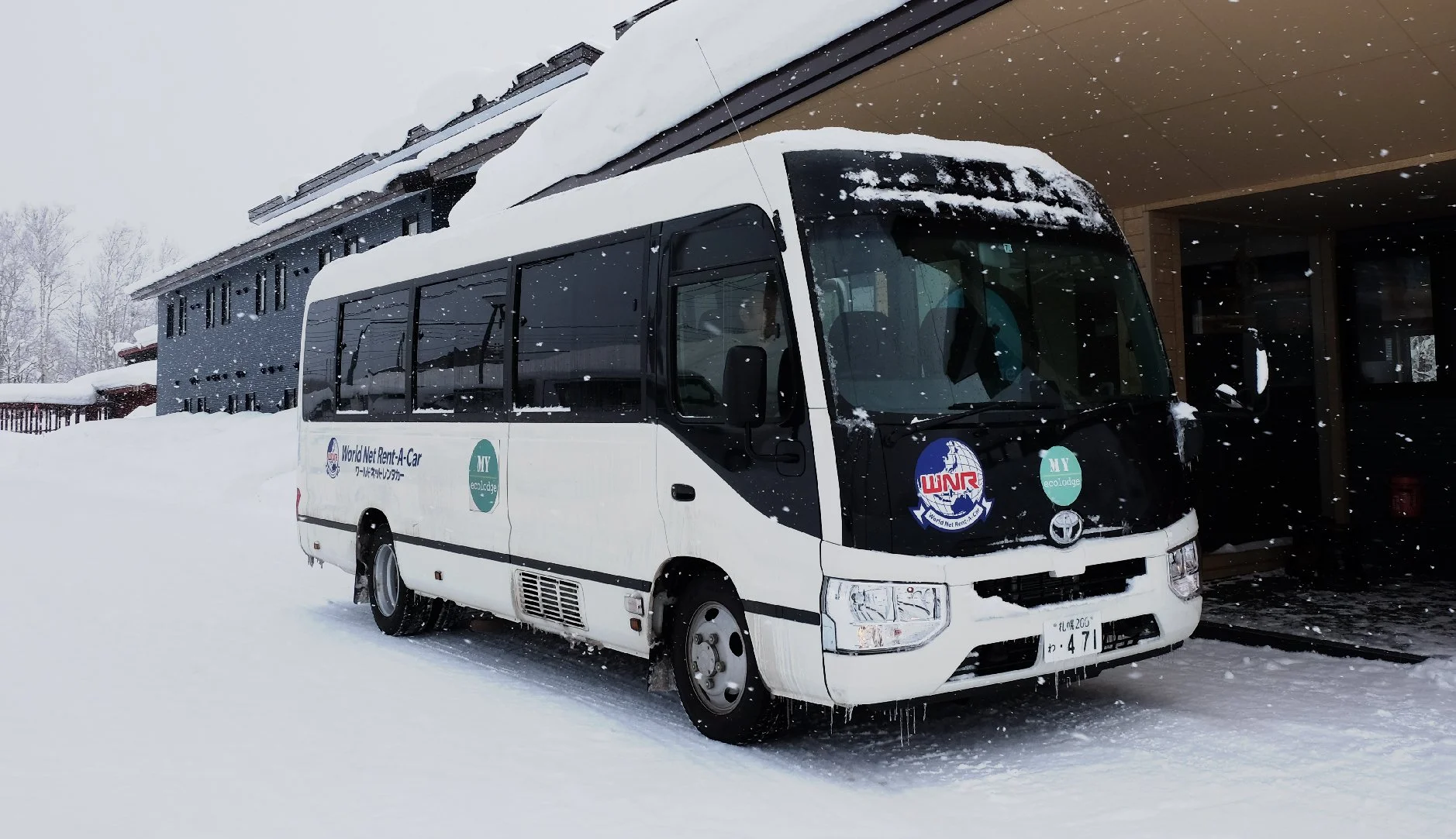 White minibus with snow on the roof, parked outside a building during snowfall, with logos and Japanese license plates visible.