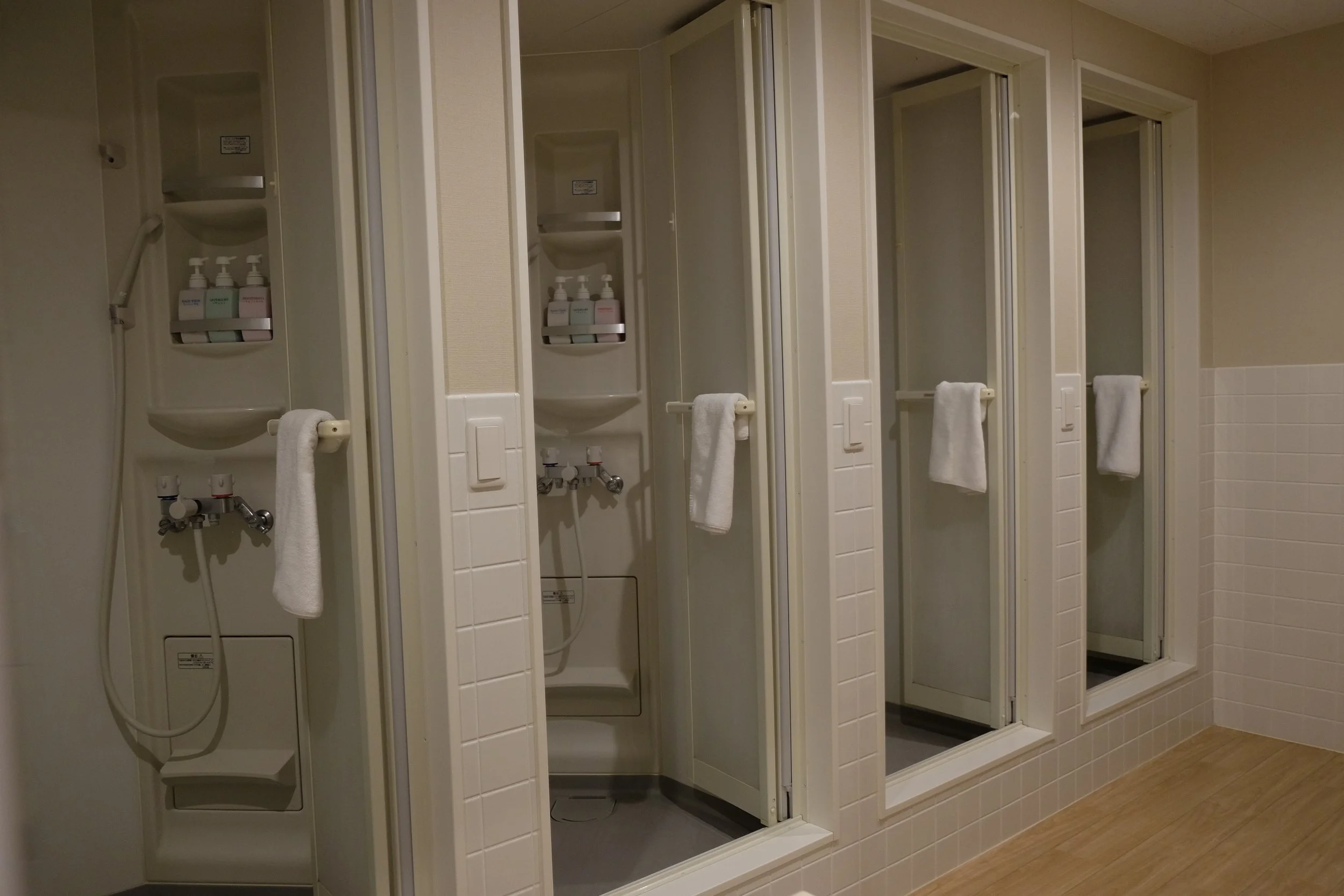 Public shower area with multiple shower stalls, each equipped with a showerhead, soap dispensers, and towel bars, set against tiled walls and wooden flooring.