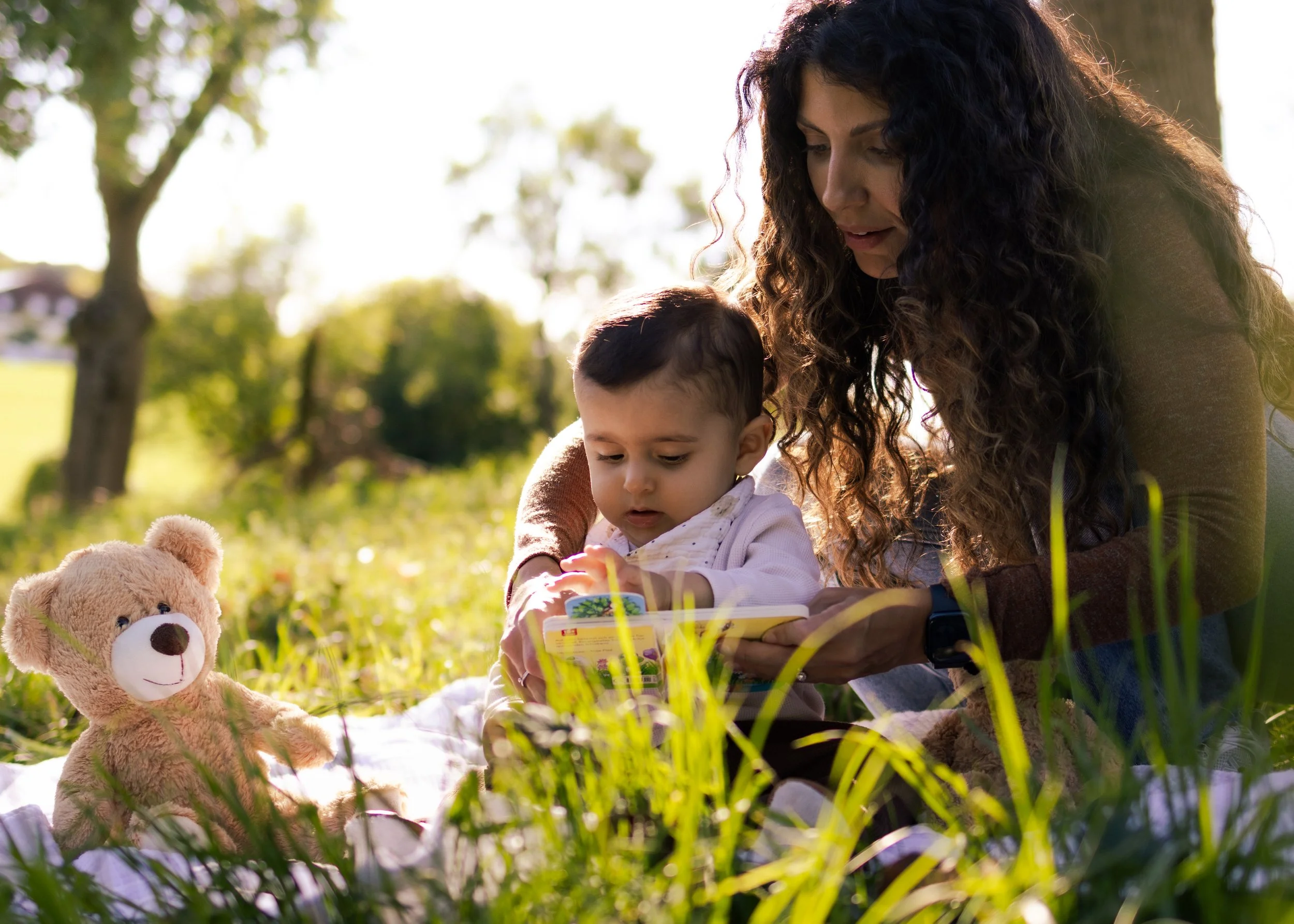 Eine Frau mit dunklen, lockigen Haaren sitzt mit einem kleinen Jungen im Freien auf dem Gras, liest gemeinsam ein Buch und hat einen Teddy dabei.