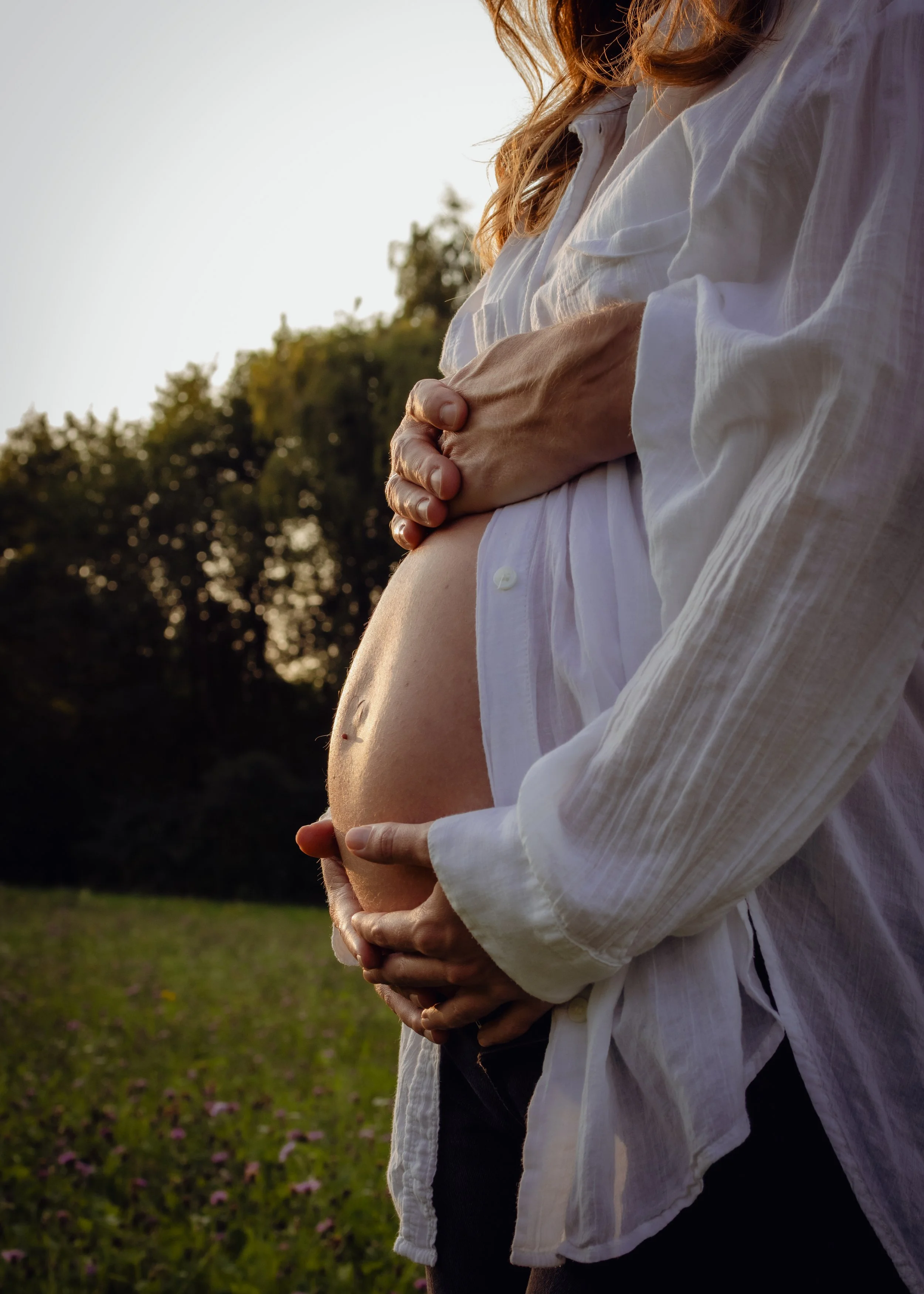 Eine schwangere Frau, die in einem weißen Hemd ist, wird im Freien bei Sonnenuntergang gehalten, während ihr Mann den Bauch umfässt.