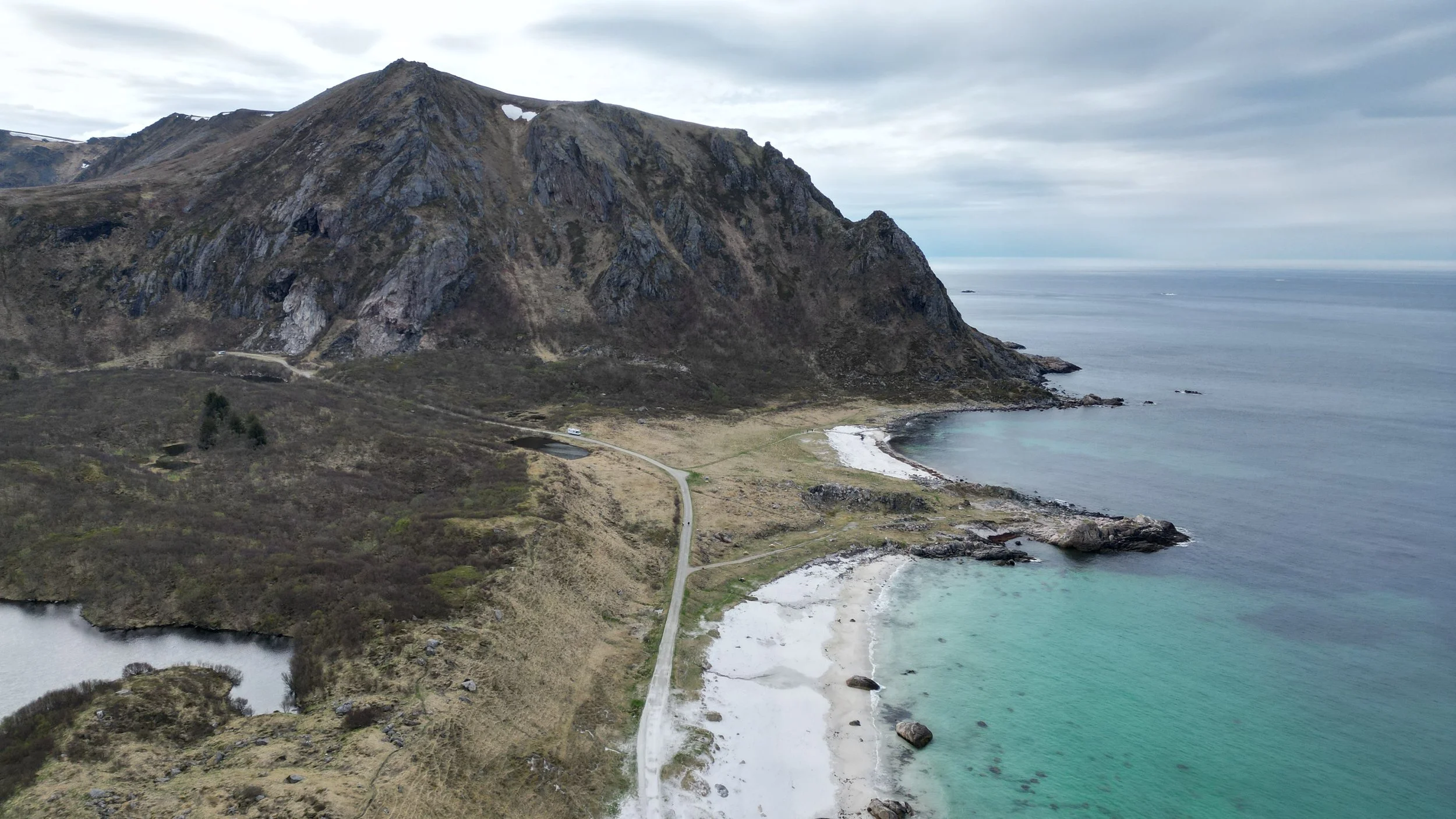 Landschaft in Norwegen mit einem großen Berg, einer Küste mit weißem Sandstrand und türkisem Wasser, bei bewölktem Himmel.