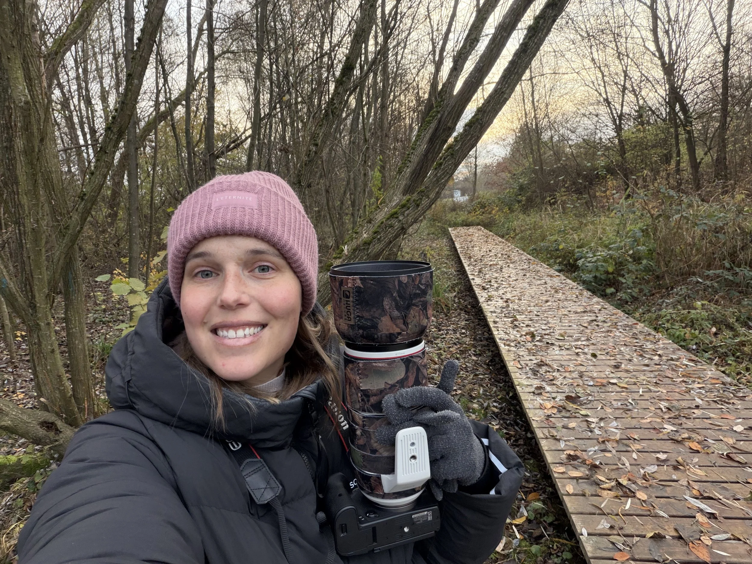 Eine lachende Frau, eine Wildtierfotografin, in wetterfester Kleidung und Mütze nimmt ein Selfie im Wald, während sie eine Kamera mit Camouflage-Muster hält, auf dem herbstlicher Laub liegt, und es ist eine Holzbrücke im Hintergrund
