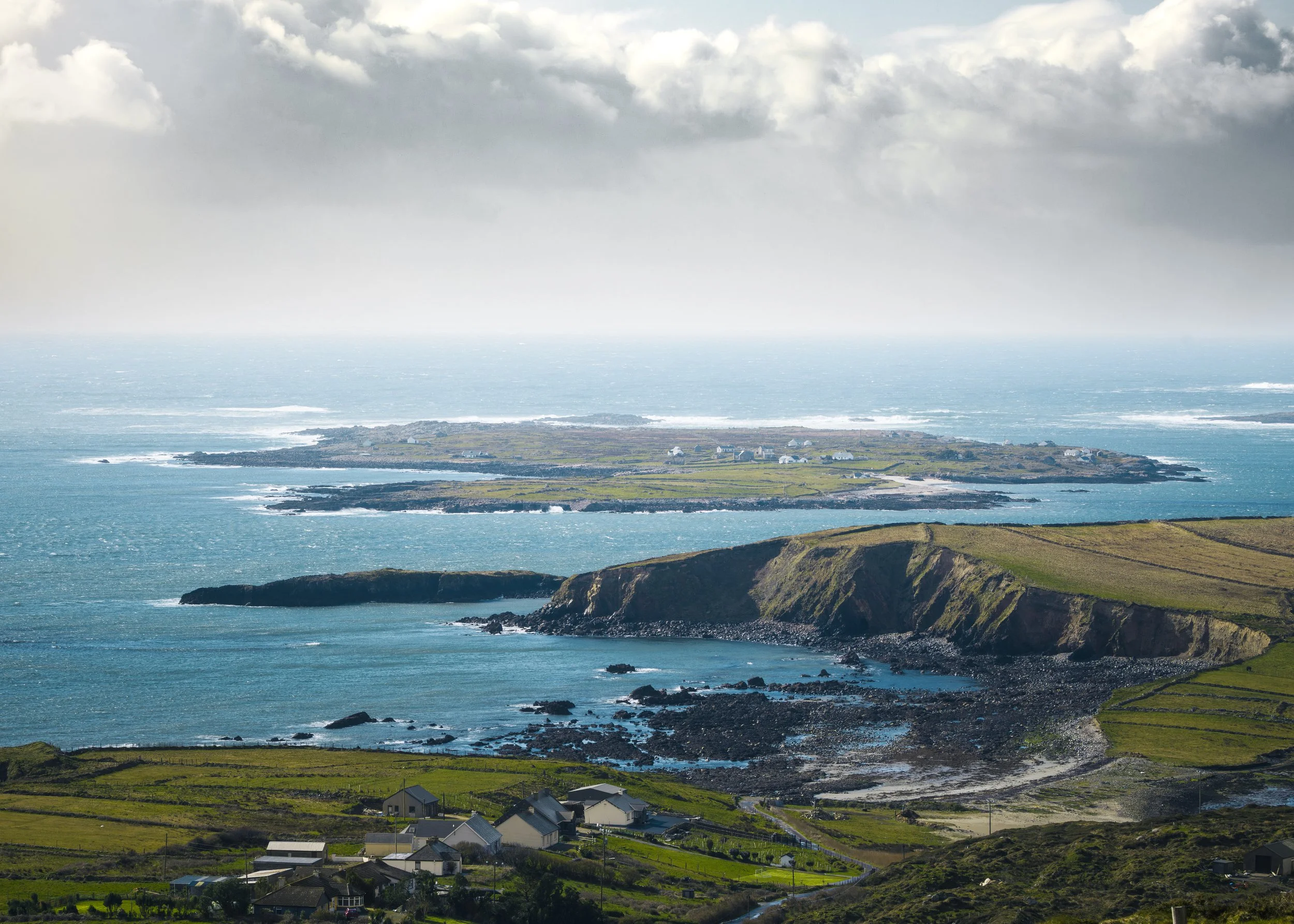 Küstenlandschaft in Irland mit grünen Hügeln, Häusern im Vordergrund, einer Insel im Meer, dunklen Wolken am Himmel und dem Ozean im Hintergrund.
