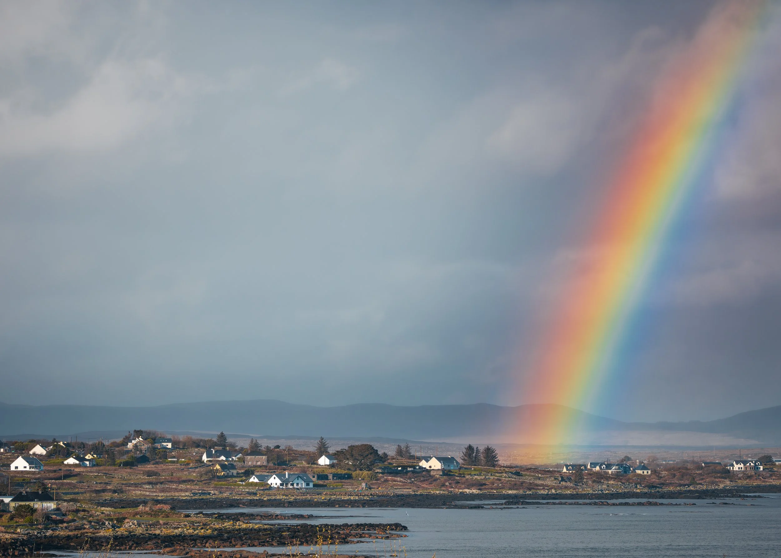 Blick auf eine Küstenregion mit mehreren weißen Häusern, dunkler Meeresküste, Berge im Hintergrund, und ein bunter Regenbogen am Himmel.