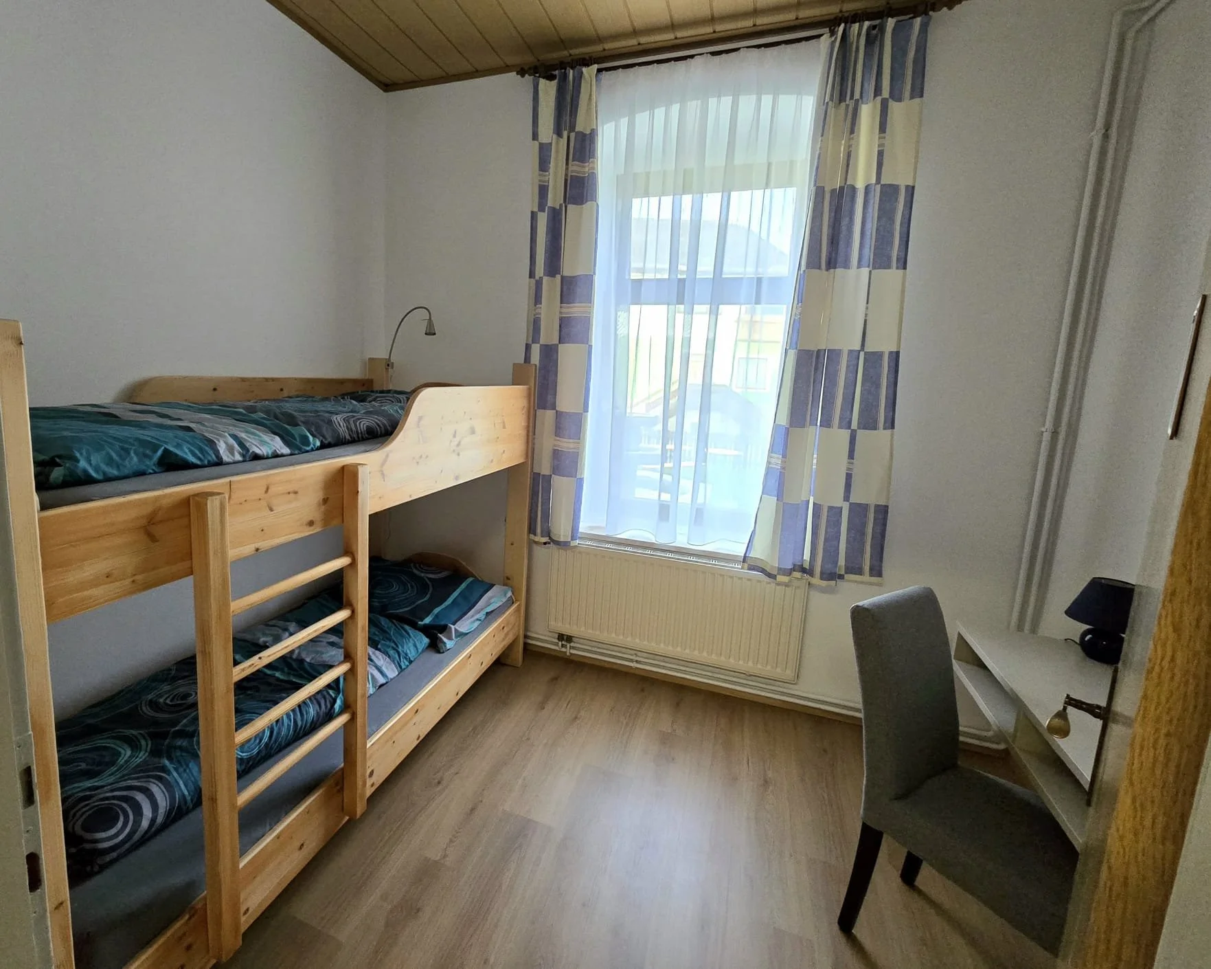 Ein Zimmer mit einem Etagenbett aus Holz, einem Fenster mit blauen und gelben Vorhängen, einem grauen Stuhl und einem kleinen weißen Schreibtisch mit einer schwarzen Tischlampe.