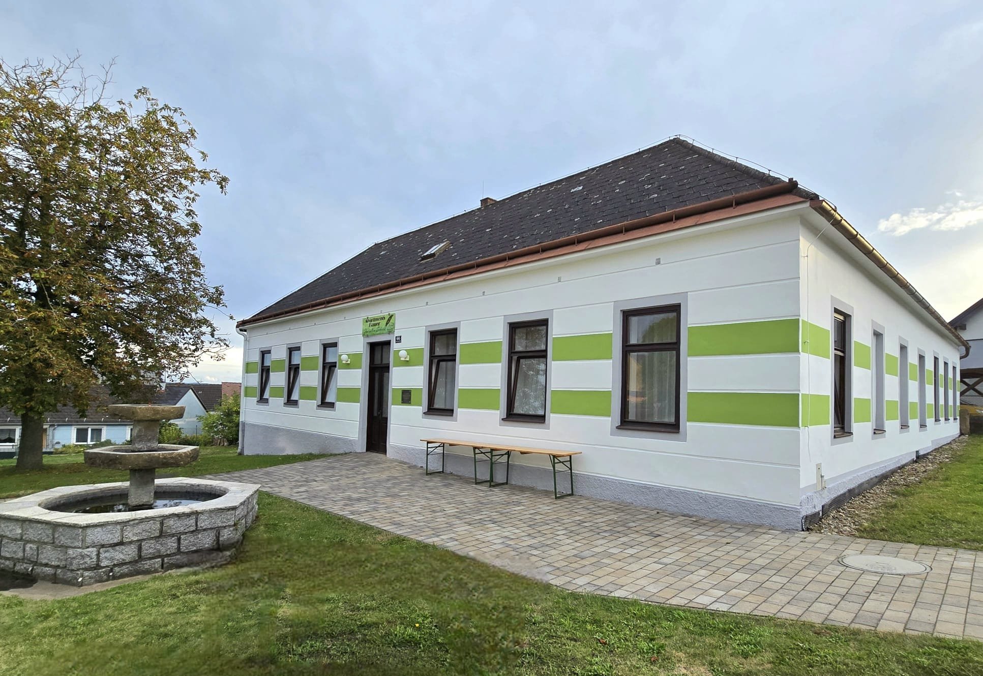 Ein weißes Gebäude mit grünen Streifen, mehrere Fenster, ein Eingang mit einer Tafel darüber, ein Garten mit einem Brunnen und einem Baum, gepflasterter Weg vor dem Haus.
