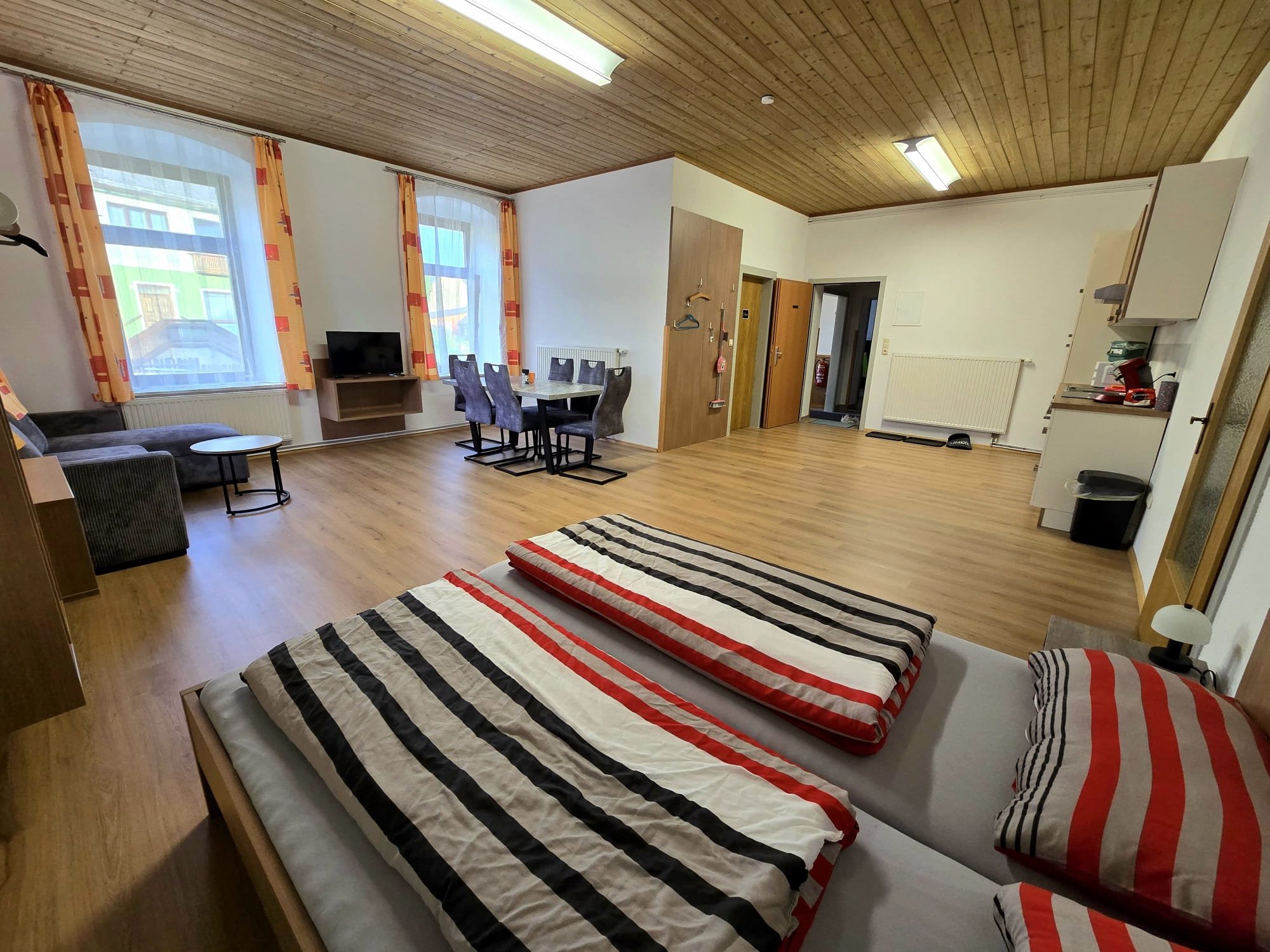 Wohnzimmer mit zwei zusammengeschobenen Betten, einem Wohnzimmerbereich mit Sofa und Fernseher, Essbereich mit Tisch und Stühlen, Holzdecken und großen Fenstern mit bunten Gardinen.