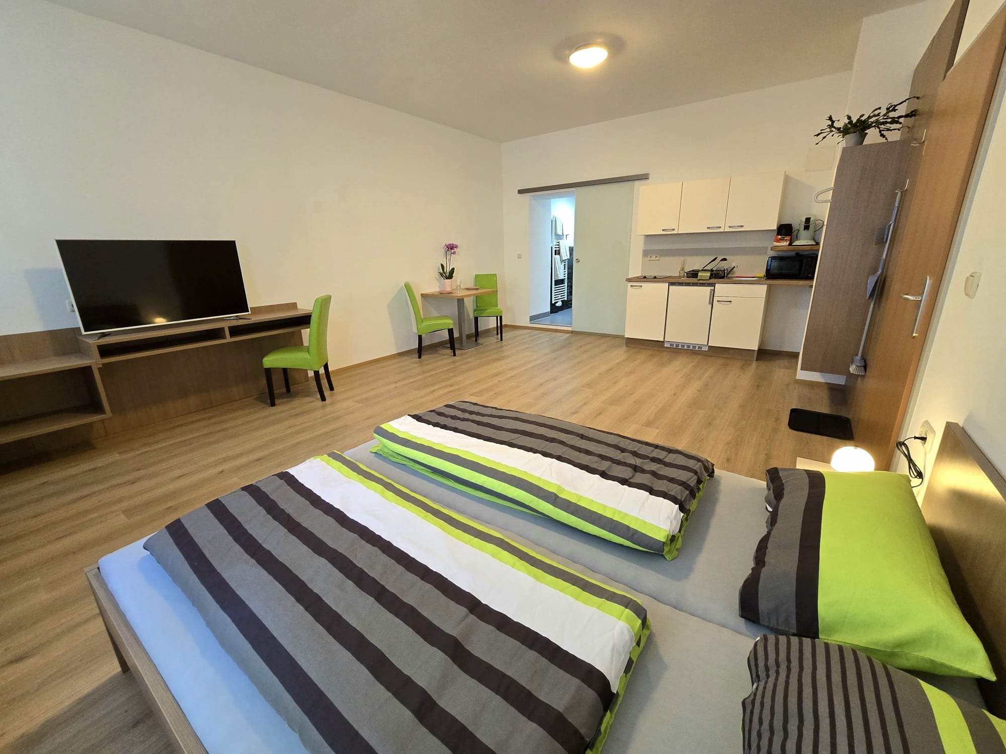 Ein Schlafzimmer mit einem Doppelbett, das mit grün, weiß, grau und braun gestreiften Bettdecken und Kissen bedeckt ist. Im Hintergrund befindet sich eine kleine Sitzecke mit einem Tisch und zwei grünen Stühlen, sowie eine offene Küche mit weißen Schränken und Geräten.