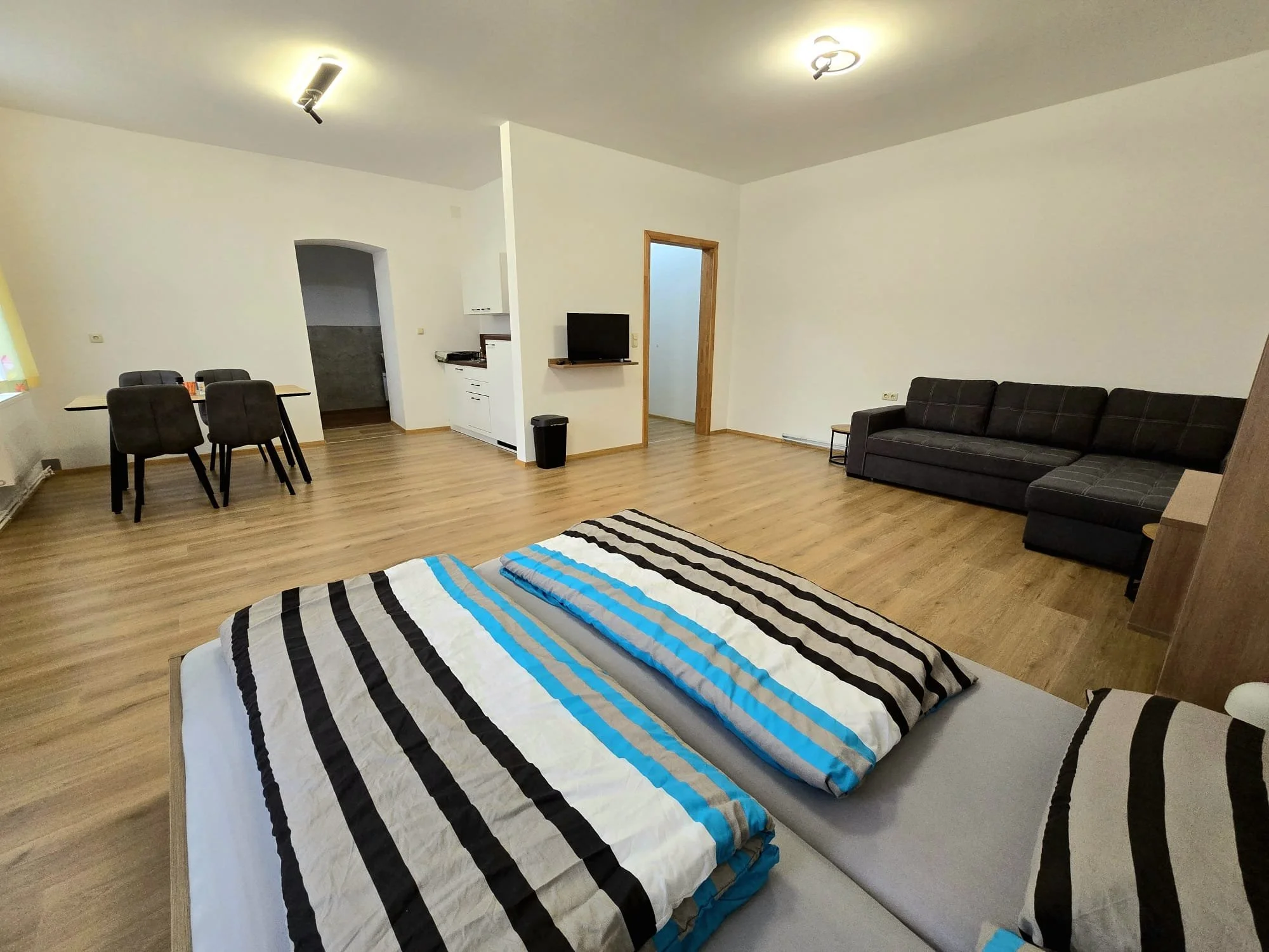 Großes Wohnzimmer mit Holzfußboden, Doppelbett, schwarzer Couch, Fernseher, Esstisch mit vier Stühlen, weiße Wände, zwei Deckenleuchten, offener Küchenbereich im Hintergrund.
