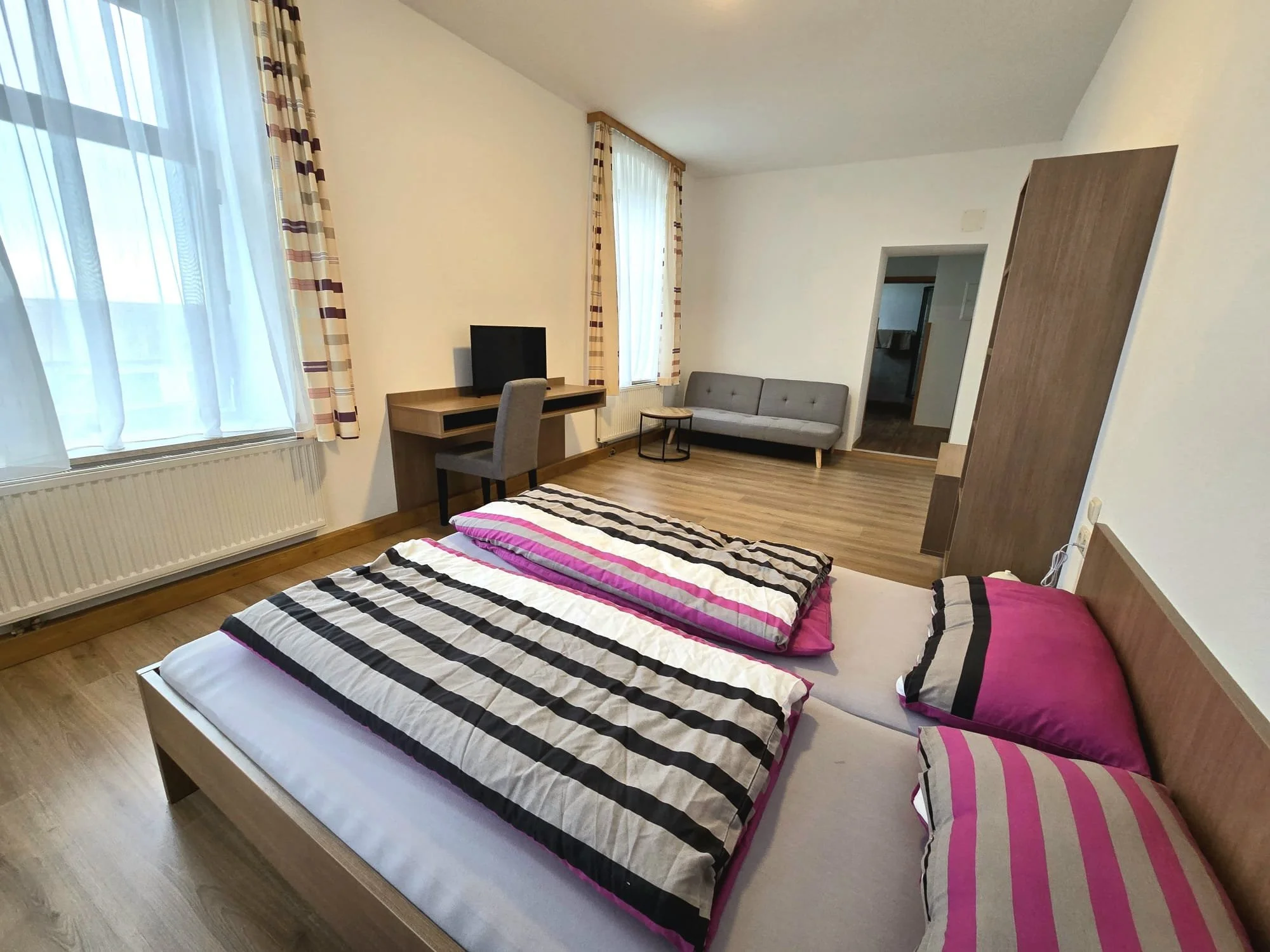 Ein Schlafzimmer mit einem Doppelbett, das mit schwarz-weiß gestreiften Bettwäsche und zwei Kissen in Pink- und Streifenmustern bedeckt ist, großen Fenstern mit weißen Vorhängen und plaidfarbenen Gardinen, einem kleinen Schreibtisch mit einem Monitor und Stuhl, einem grauen Sofa, einem kleineren Tisch, einem Kleiderschrank und einem Eingang zu einem anderen Raum.