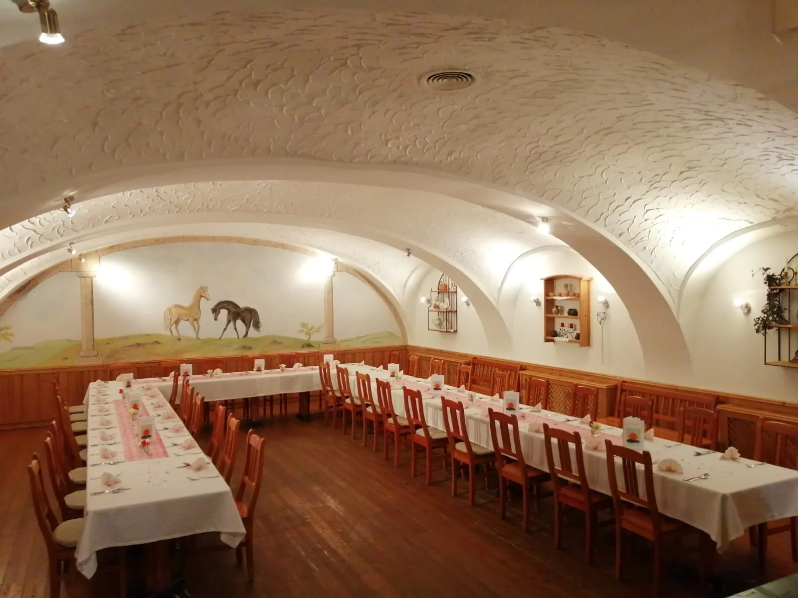 Ein festlich gedeckter Speisesaal mit langen Tischen, weißen Tischdecken, kleinen Blumenvasen und Holzstühlen. Die Wände sind weiß mit Wandmalereien von Pferden und Landschaften, und das Deckeninnere ist gewölbt und verziert.