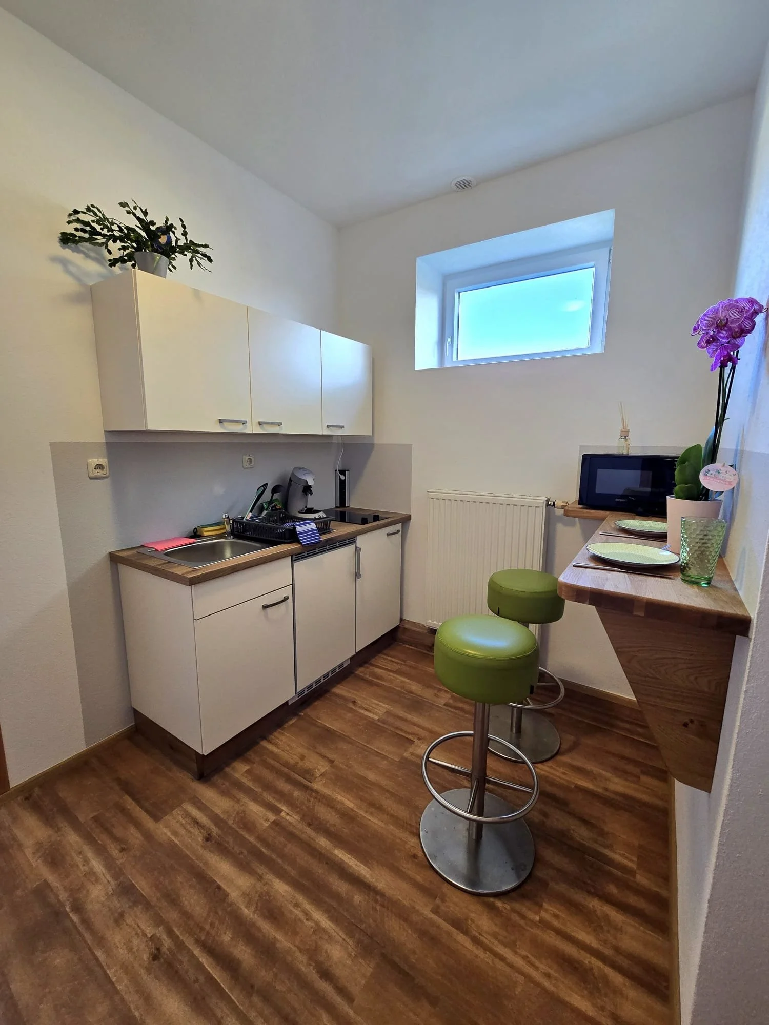 Kleine Küche mit weißen Schränken, dunkelbraunem Holzfußboden, einem Fenster, einer Fensterbank mit Vase und Orchidee, sowie einem kleinen hohen Tisch mit grünem Stuhl.