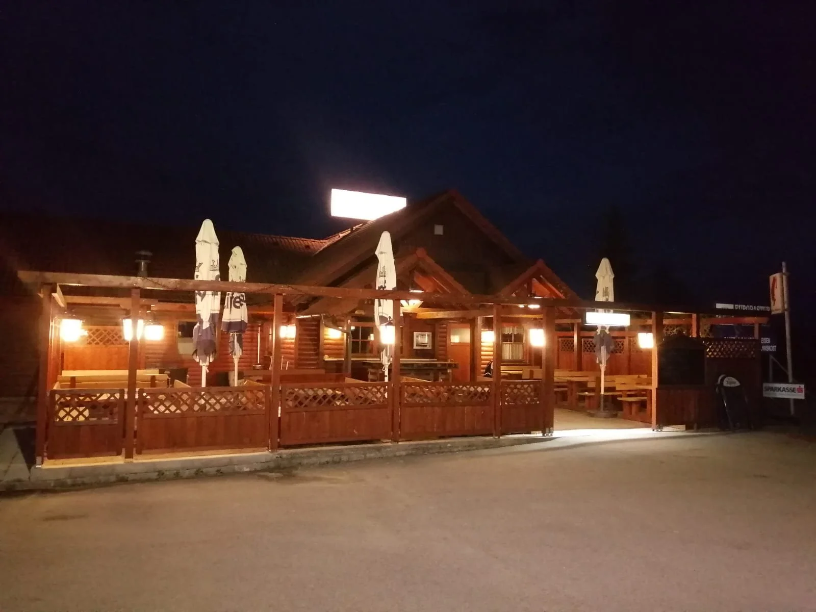 Ein Restaurant mit Holzkonstruktion und Außenbereich bei Nacht, beleuchtet mit warmem Licht, mit mehreren geschlossenen Sonnenschirmen.