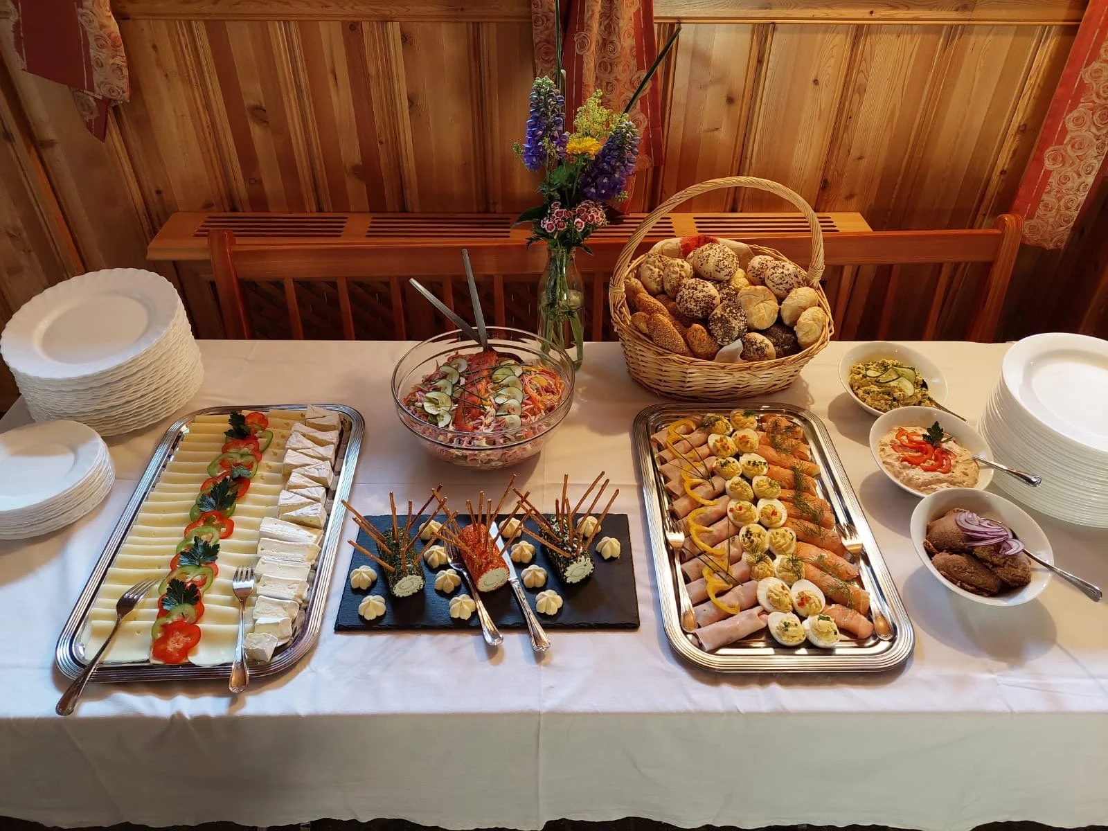 Buffet mit verschiedenen kalten Speisen, darunter Käse, Wurst, Gemüse, Kartoffeln, ein Salat, eine Schale mit Kaviar und eine Planke mit belegten Broten. Es gibt auch einen Korb mit Brötchen und eine Vase mit Blumen.