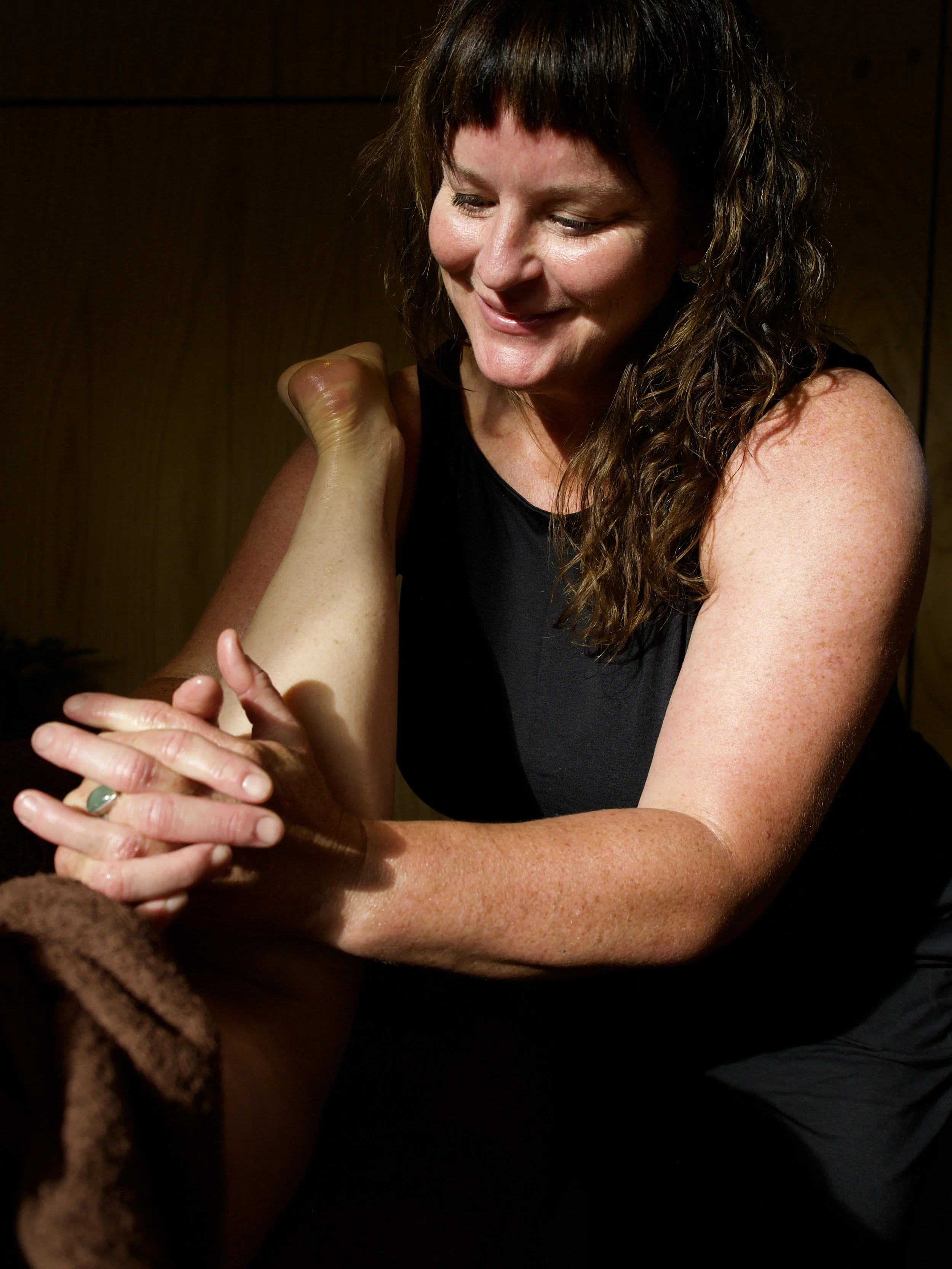 A woman with curly brown hair smiling while receiving a massage on her arm in a dimly lit room.