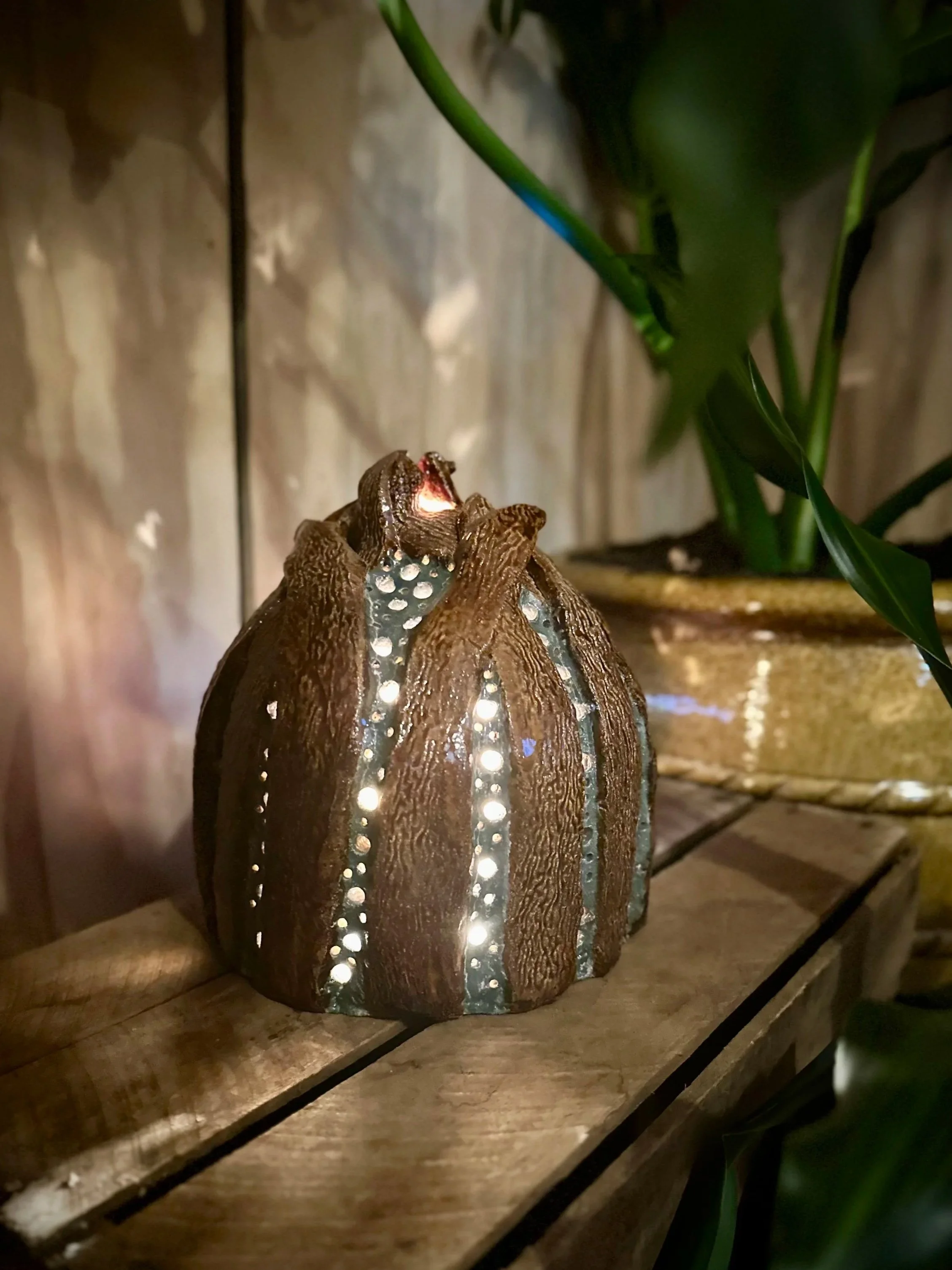 A decorative lamp shaped like a seed with illuminated small holes, placed on a wooden surface with faint green leaves and a wooden wall in the background.