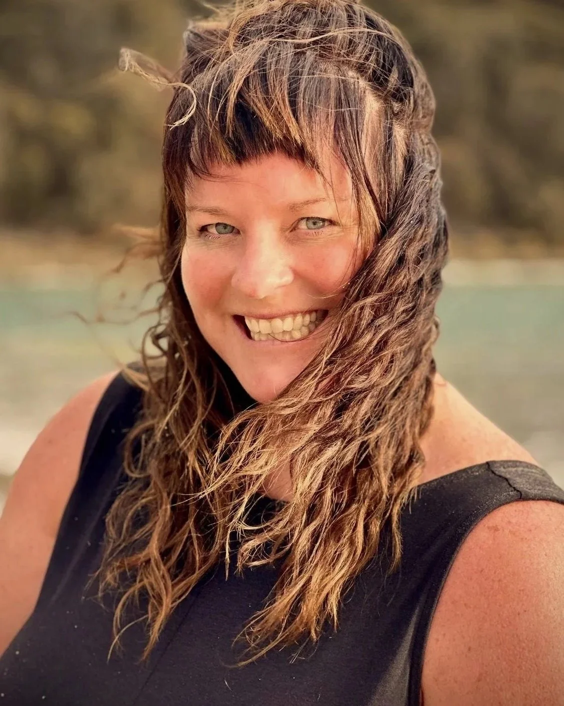Close-up of smiling woman with wet, curly brown hair, wearing a black sleeveless top, outdoors with blurred background.