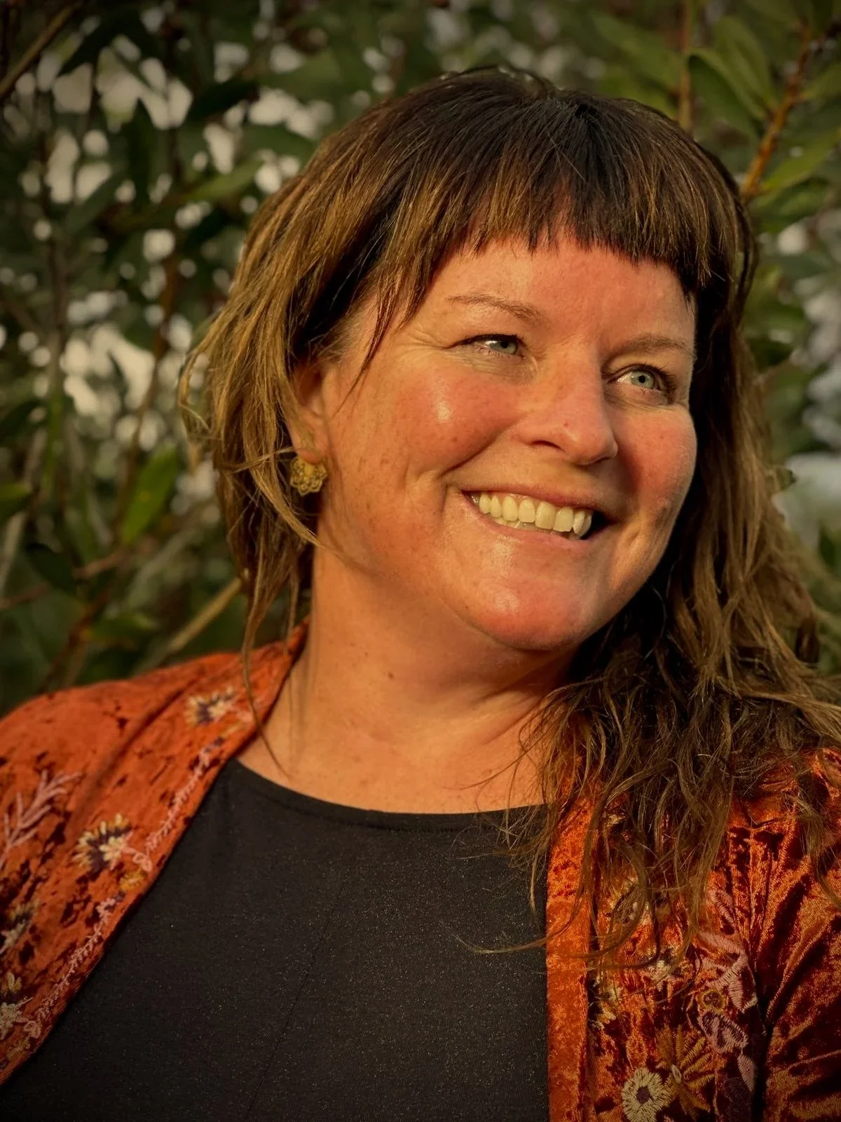 A woman with light skin and blue eyes, smiling outdoors, wearing a black top and an orange, patterned jacket.