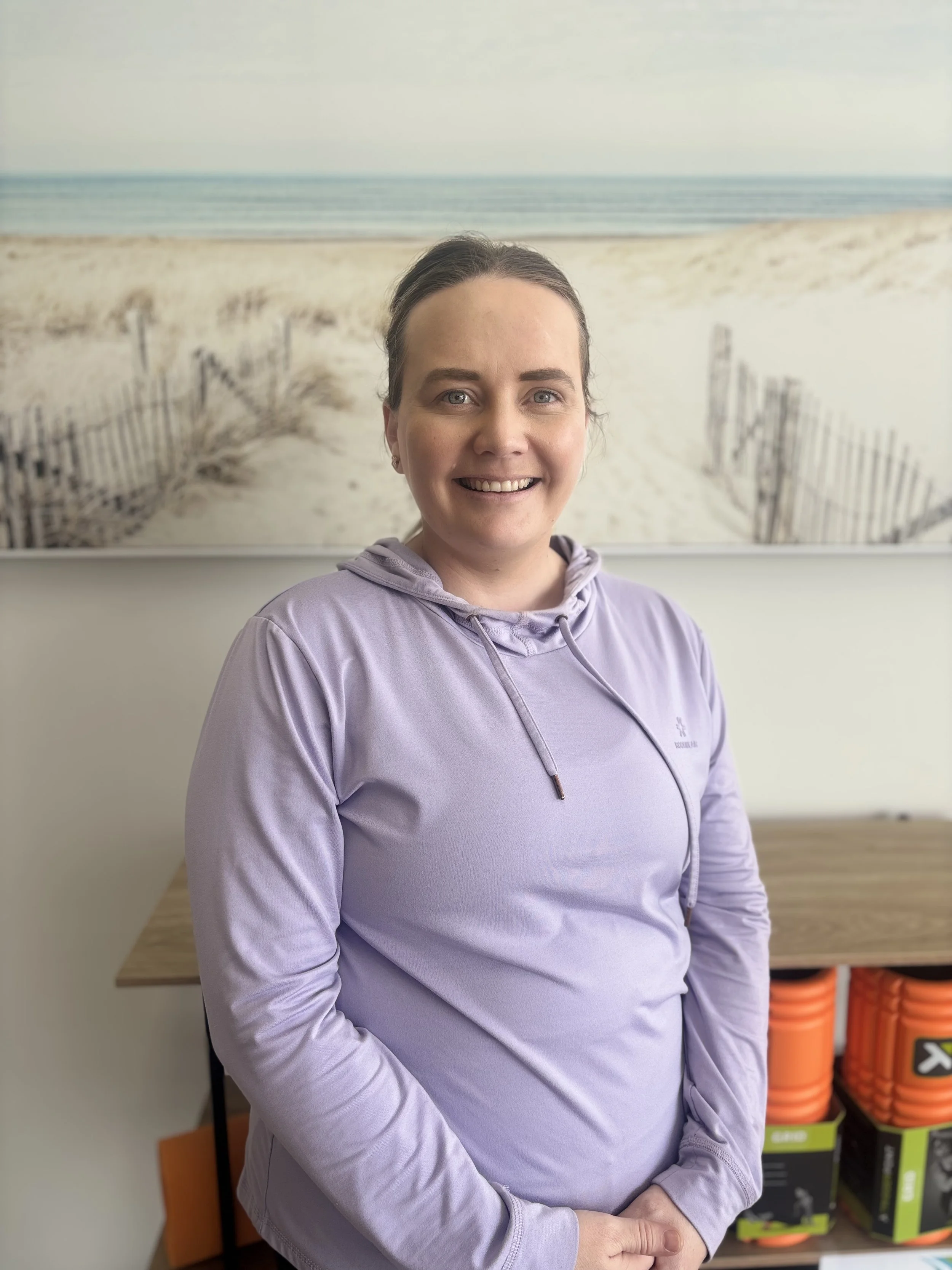 A woman standing indoors in front of a large beach-themed photograph, wearing a light purple hoodie and smiling.