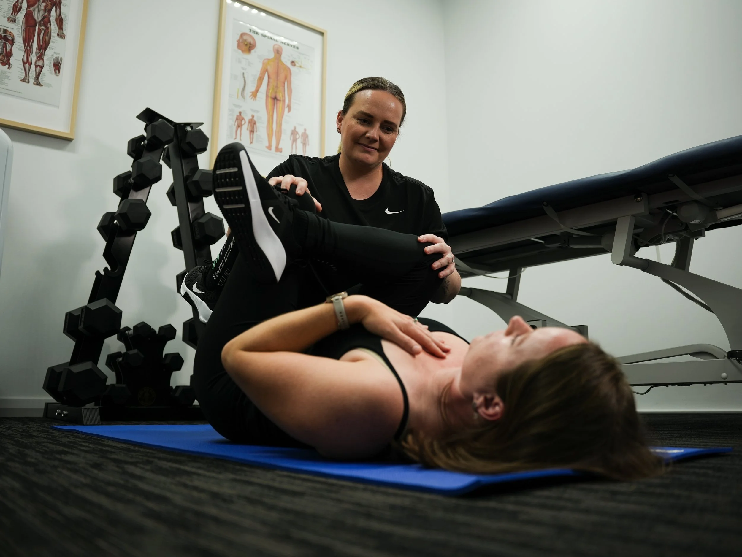 A woman lies on a blue exercise mat on the floor, holding her knee with one hand. A healthcare or fitness trainer kneels beside her, supporting her leg and observing. The setting appears to be a physical therapy or gym room, with anatomical posters on the wall and exercise equipment nearby.