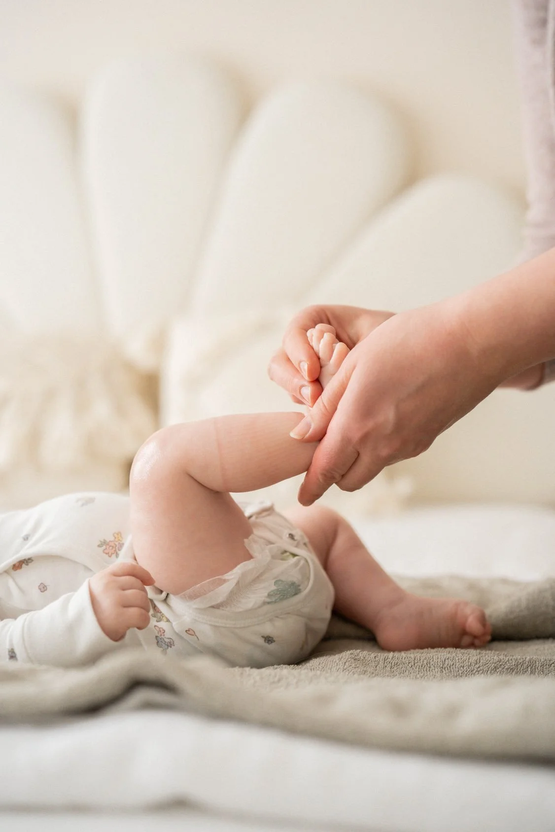 Close-up of an adult holding a baby's foot, baby lying on bed with patterned pajamas, soft neutral background.