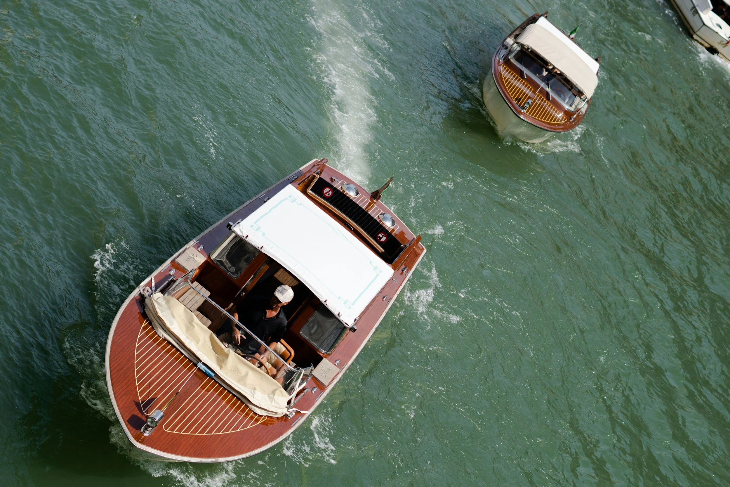Overhead view of a boat cruising on green water with a wake behind it, showing the boat's bow and partial deck with two people on board.