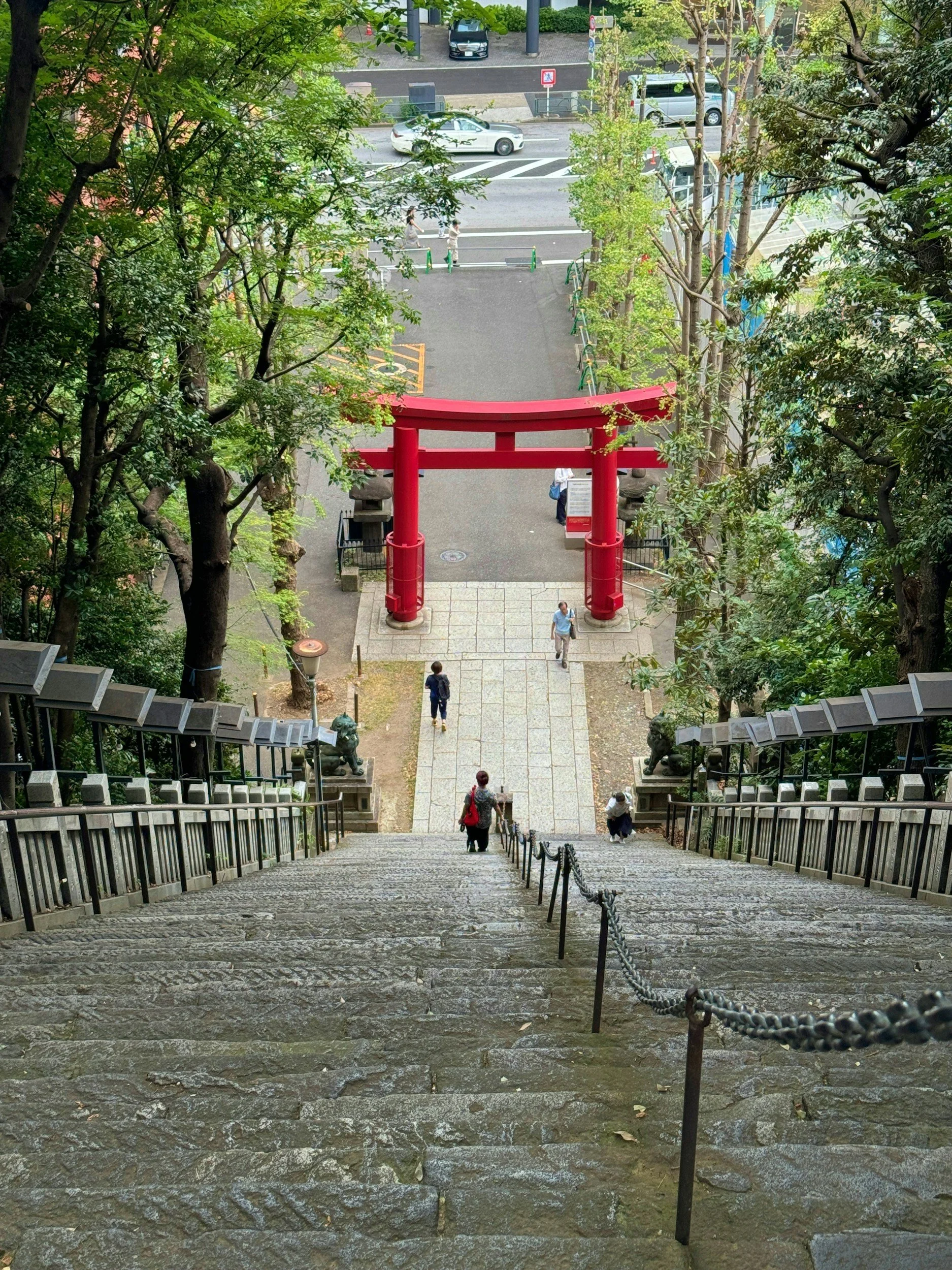Stone stairway leading down to a courtyard with a red torii gate, surrounded by trees and a sidewalk with pedestrians, in an urban area.