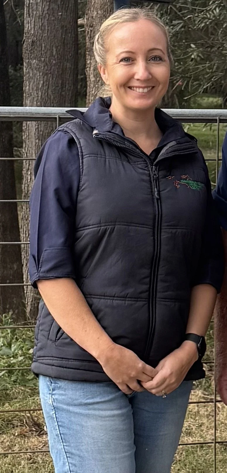 A woman with blonde hair tied back, smiling, wearing a black puffy vest with a green crocodile embroidery on the chest, blue jeans, and a black watch, standing outdoors in front of trees and a metal fence.