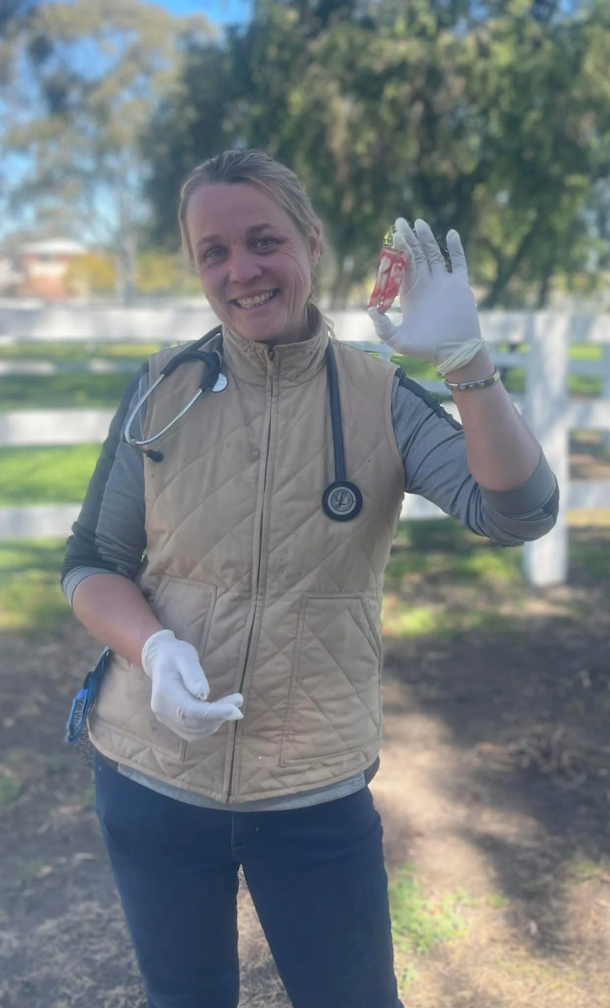A smiling woman in medical scrubs and gloves holding a small medical sample with a backdrop of trees and a white fence.