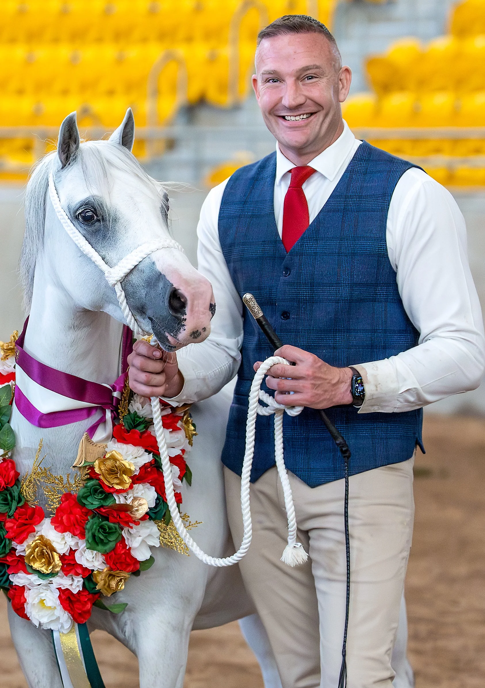 A man smiling and holding a show stick standing next to a decorated white horse with a floral and ribbon decoration, in an indoor arena with yellow seating in the background.