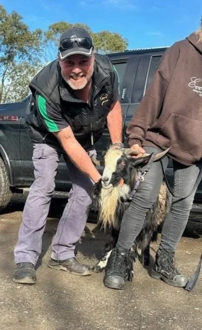 A man smiling and holding a small goat with long white beard in front of a black vehicle outdoors on a sunny day.