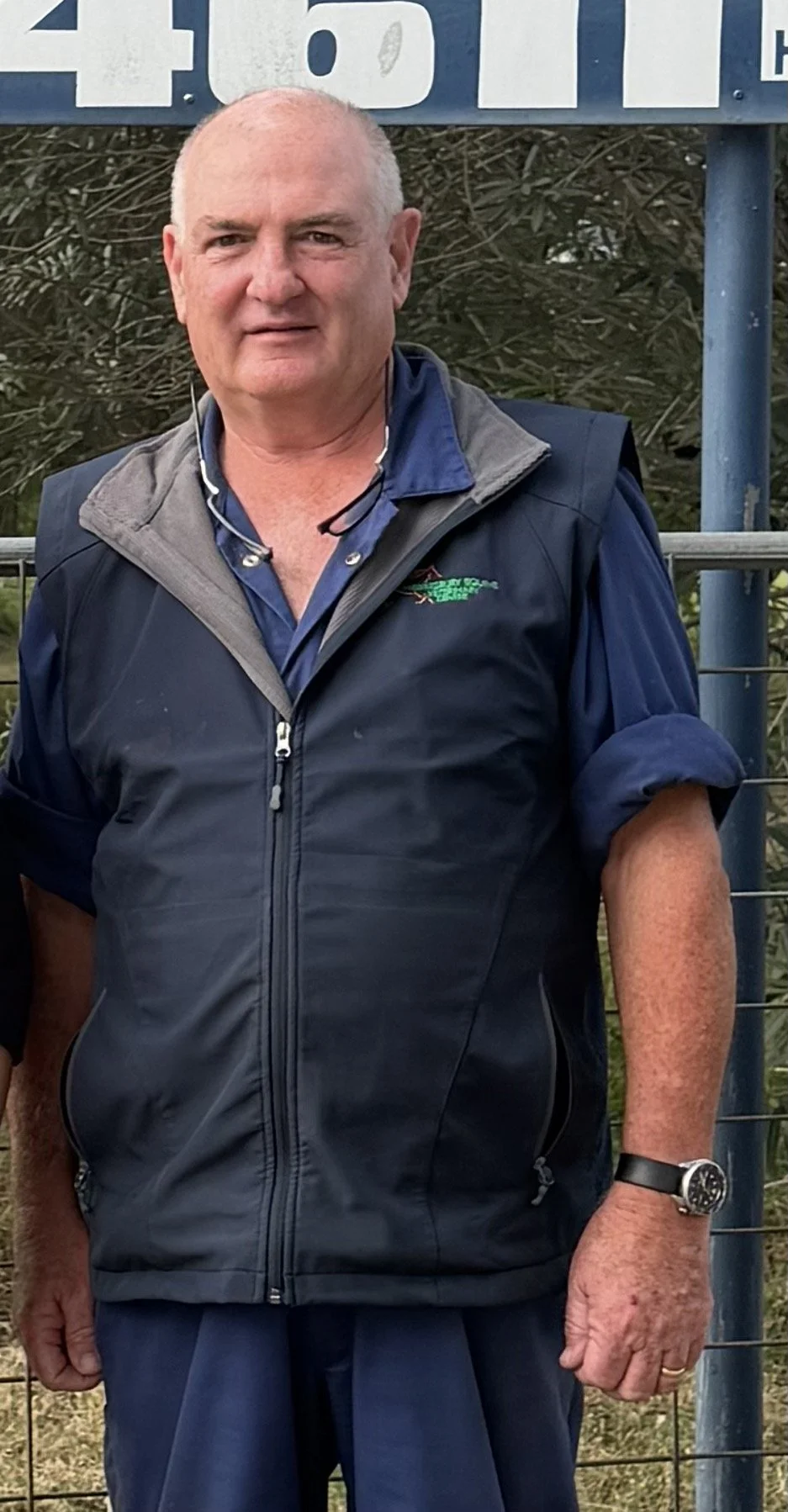 A middle-aged man with short white hair standing outdoors, wearing a navy blue windbreaker, a blue collared shirt, a wristwatch, and a necklace. There is a fence and a sign with partially visible text behind him.