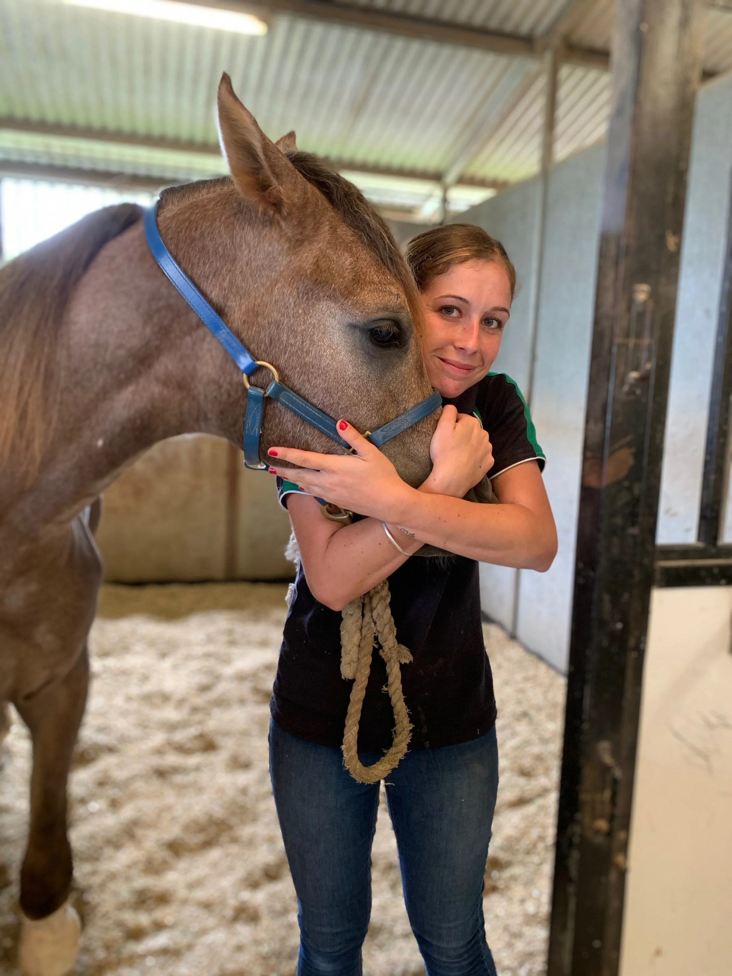 A woman hugging a light brown horse inside a stable.