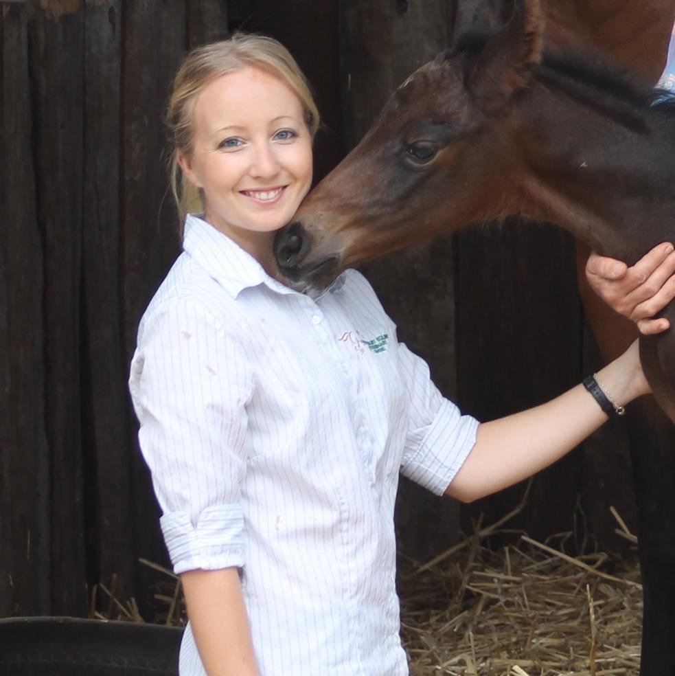A young woman with blonde hair smiling while holding a brown horse's face close to hers. They are in a wooden stable.