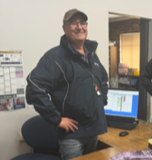 Man wearing a navy jacket and baseball cap standing in front of a computer in an office