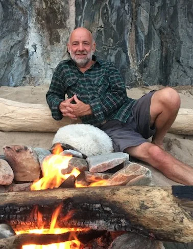A man sitting beside a small fire outdoors, with a white sheepskin beside him. and a rocky cliff in the background.