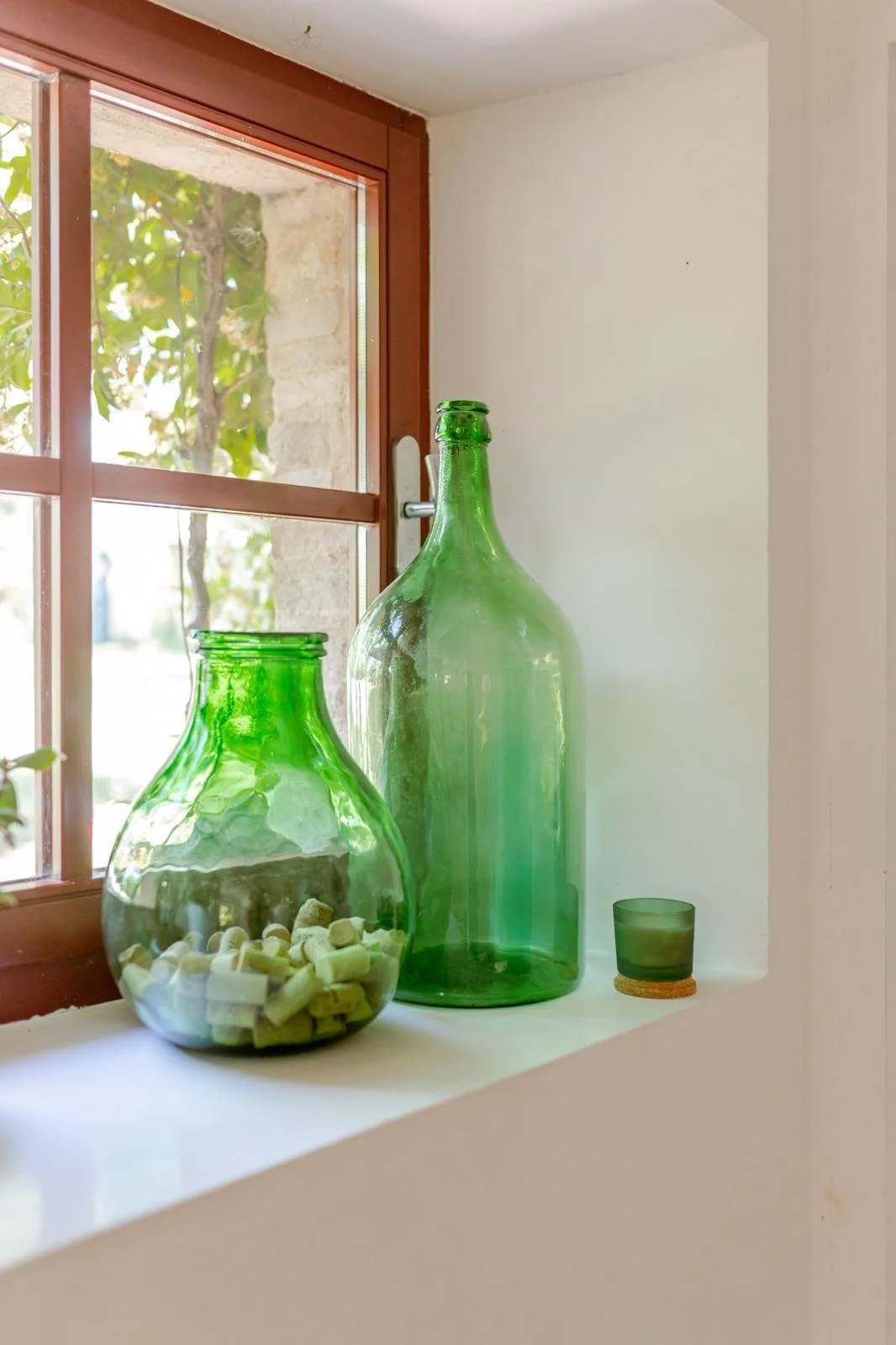 Two green glass bottles and a green glass tumbler on a white windowsill beside a window with a view of green leaves outside.