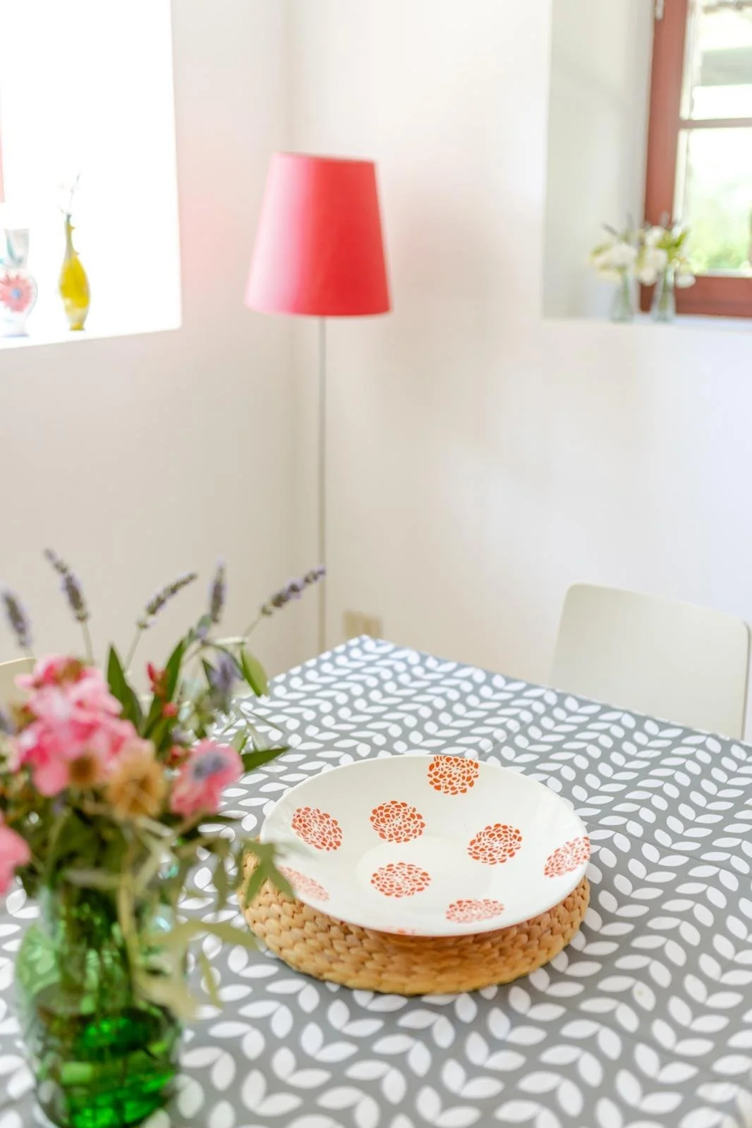 A dining table with a black and white patterned tablecloth, a white bowl with orange floral design on a woven base, a green glass vase with pink and purple flowers, a pink standing lamp, and two windows with vases of white flowers.